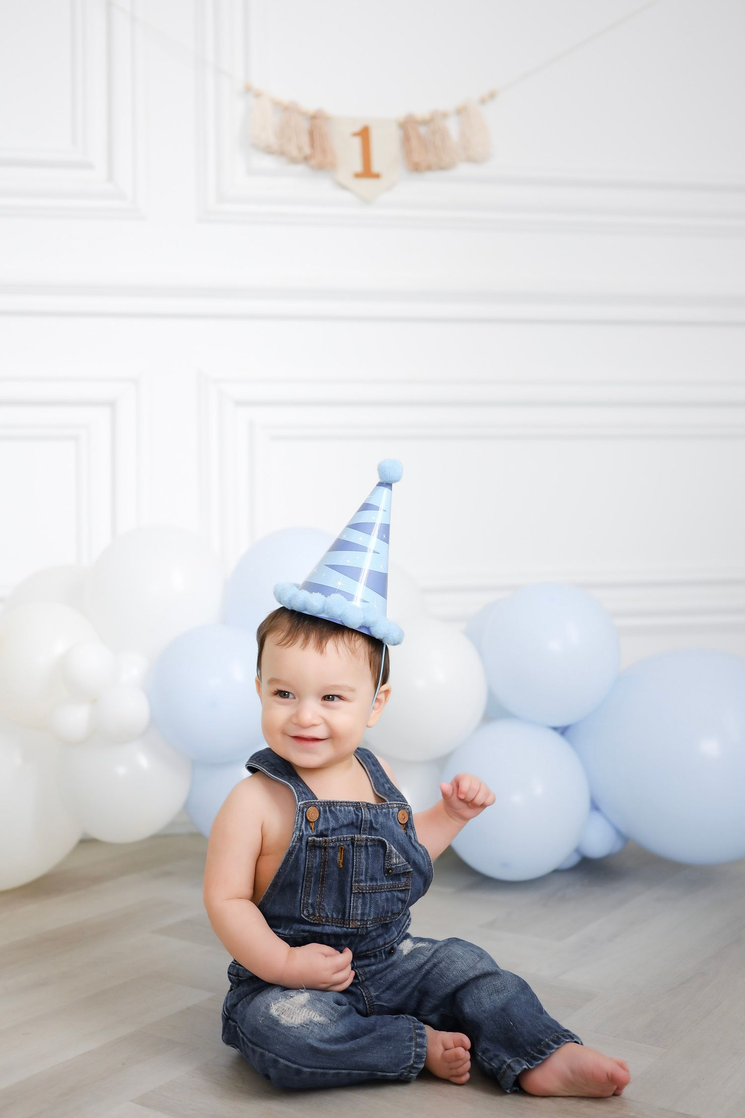Smiling baby in denim overalls and blue party hat sits by balloons celebrating 1st birthday.