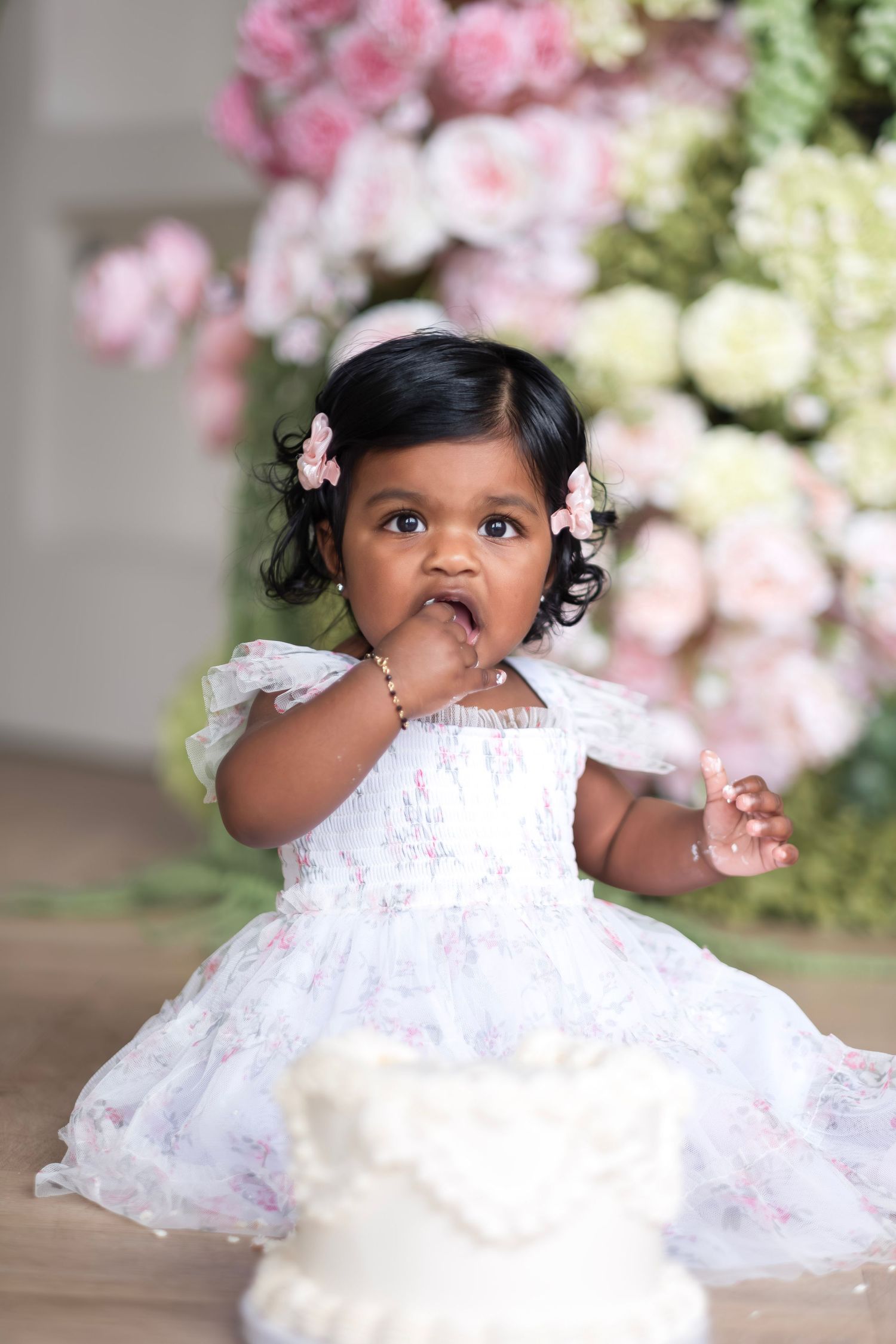 Baby girl in white floral dress eating cake at her first birthday party with pink flowers in background.