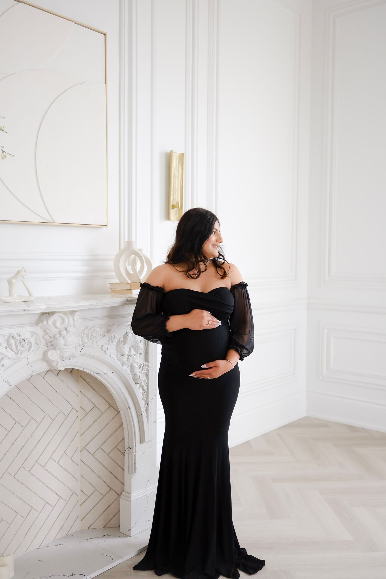 Elegant maternity photo of an expectant mother in a black off-shoulder gown standing by a white fireplace in luxurious room.