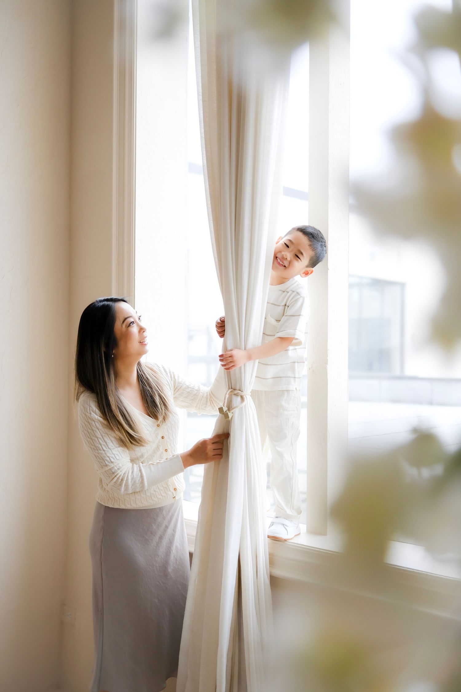 Two people in white outfits work together to adjust sheer curtains by a bright sunlit window.