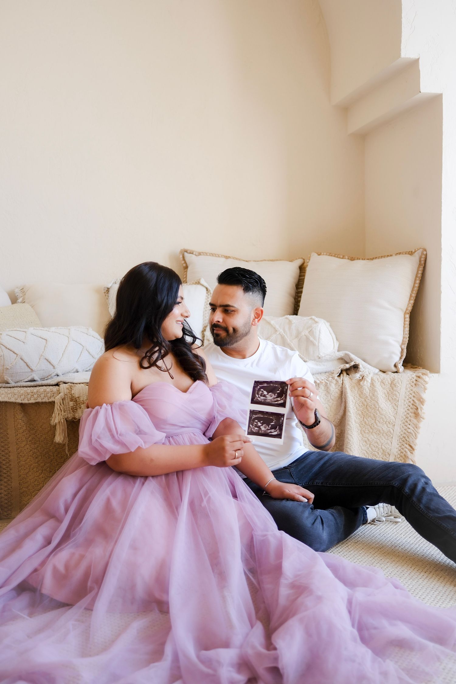 Couple in casual attire sits on bed sharing ultrasound photo while woman wears flowy pink dress.