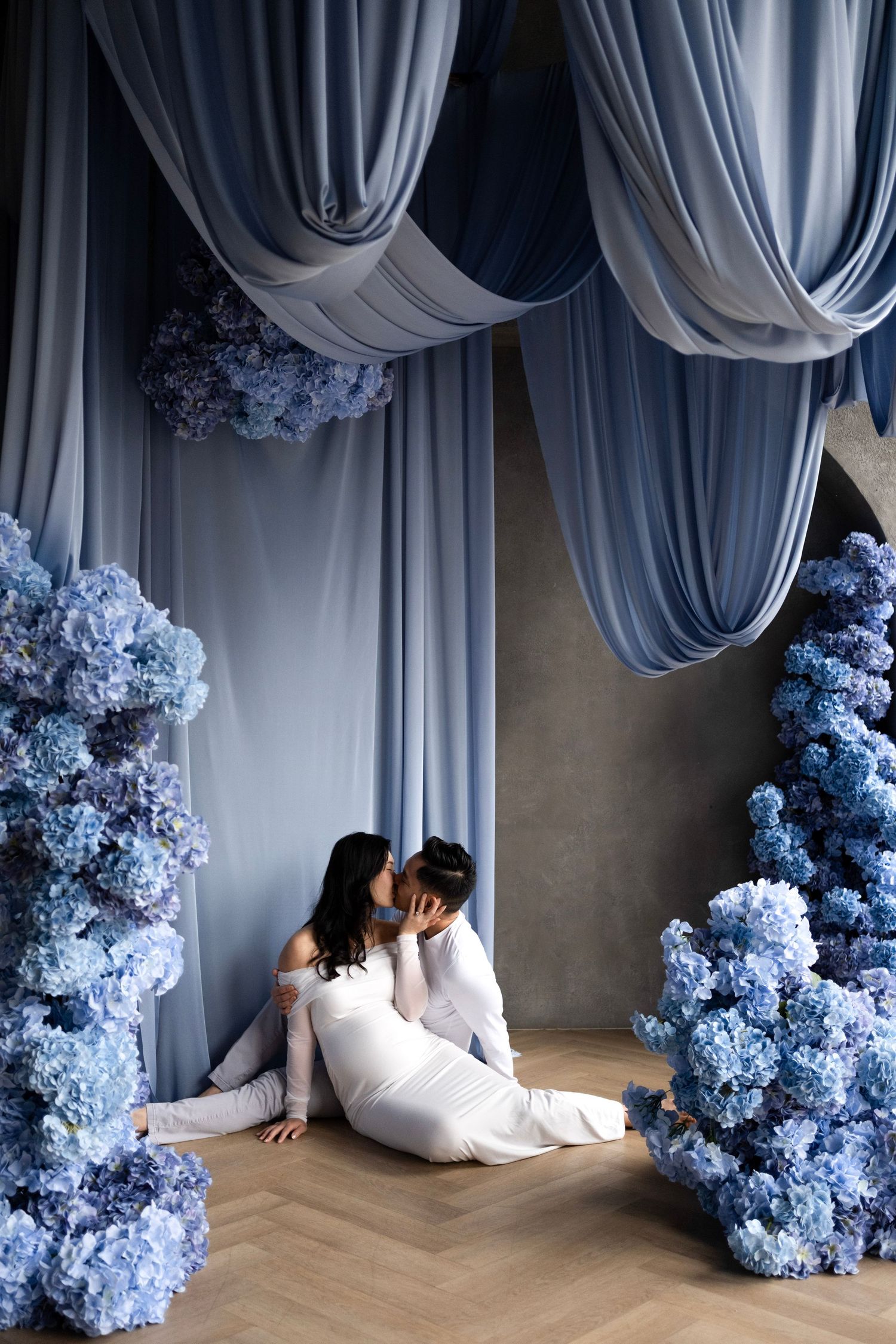 Couple in white outfits sitting among blue hydrangeas with elegant draped fabric backdrop.