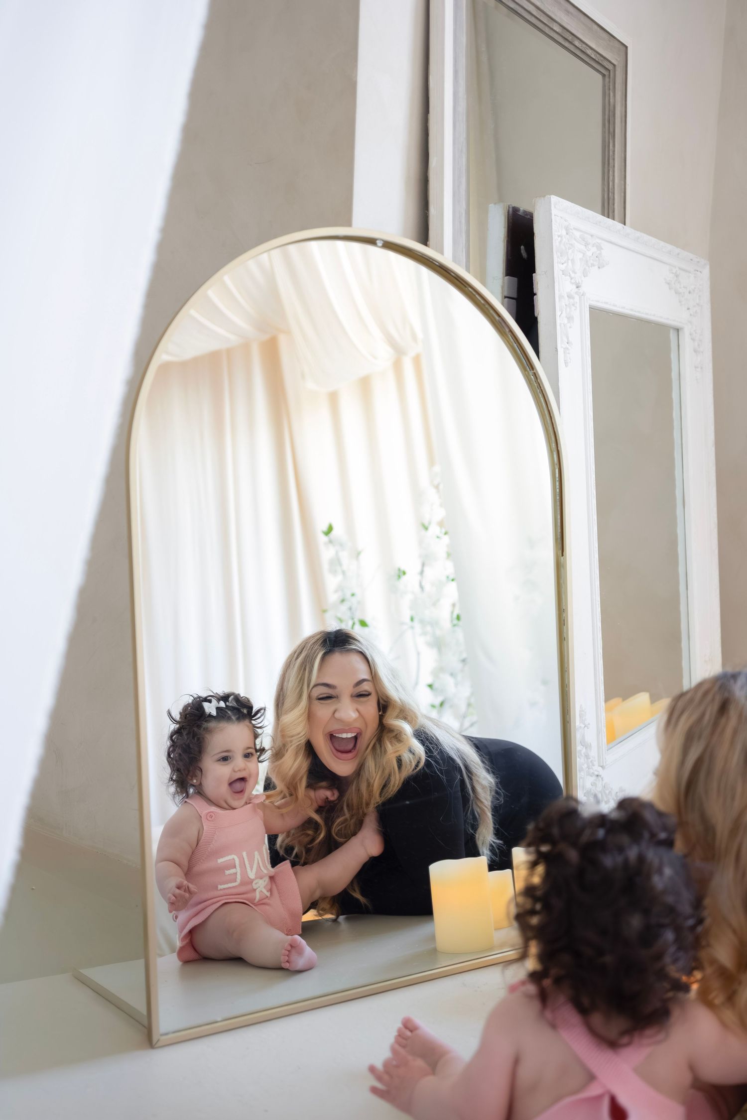 Woman and baby girl laughing together reflected in gold arch mirror in bright white room.