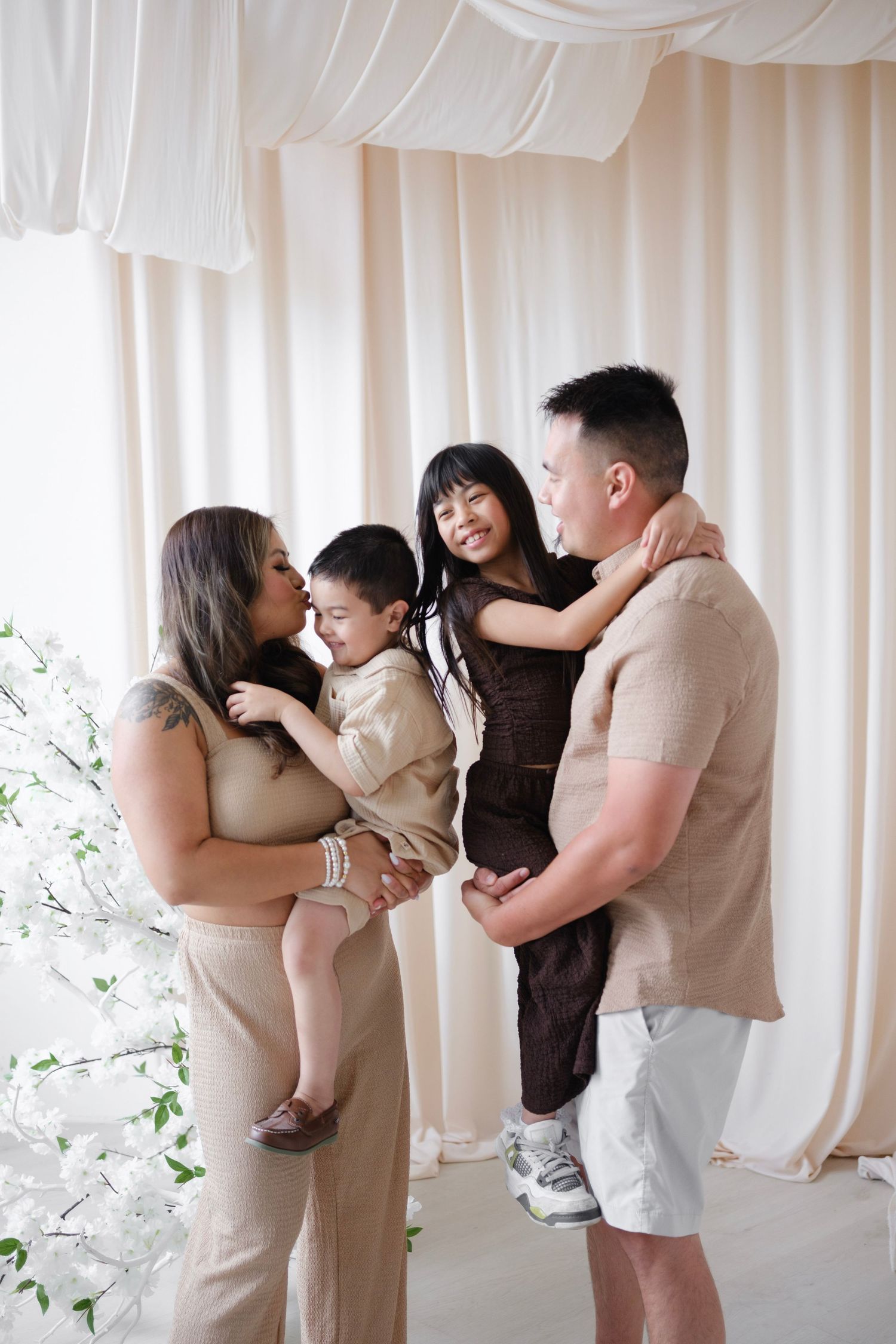 A happy family of four poses together in a bright studio with white drapes and floral decor.
