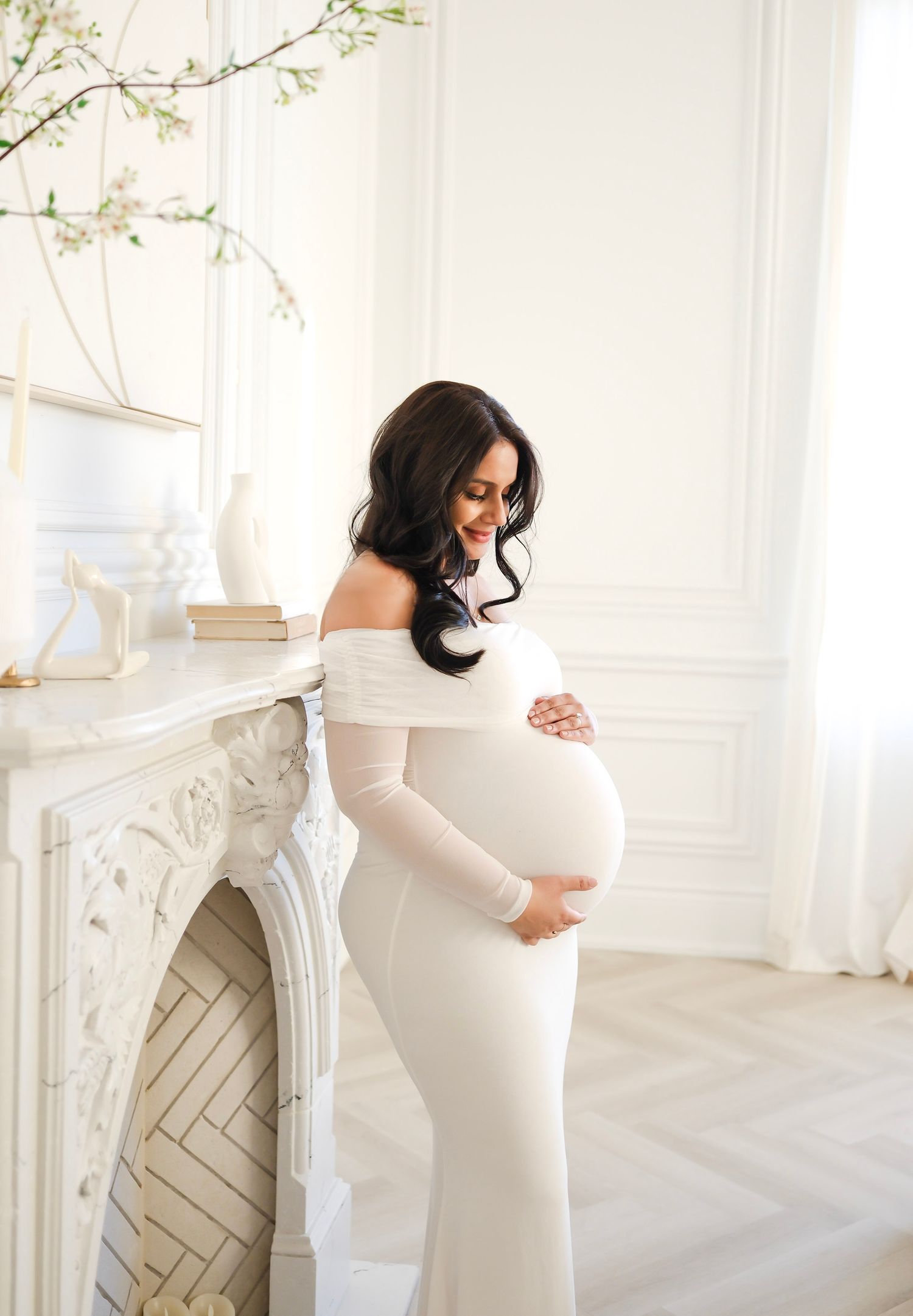 Elegant maternity portrait in a white off-shoulder gown standing by an ornate fireplace in a bright, airy room.