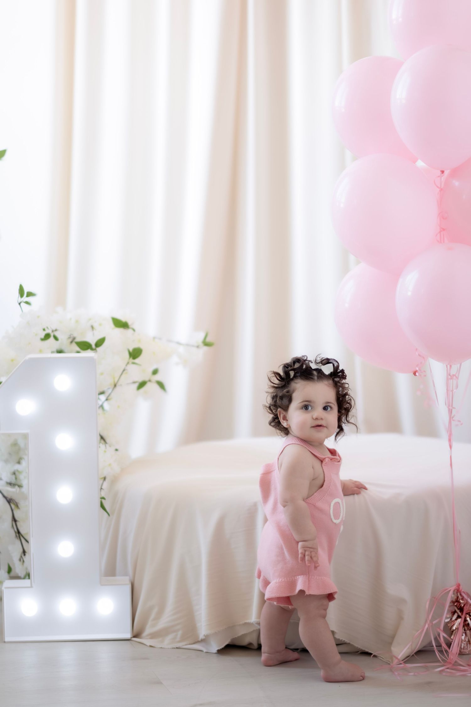 A baby girl in a pink dress stands next to pink balloons and a lit number 1 sign for her first birthday.