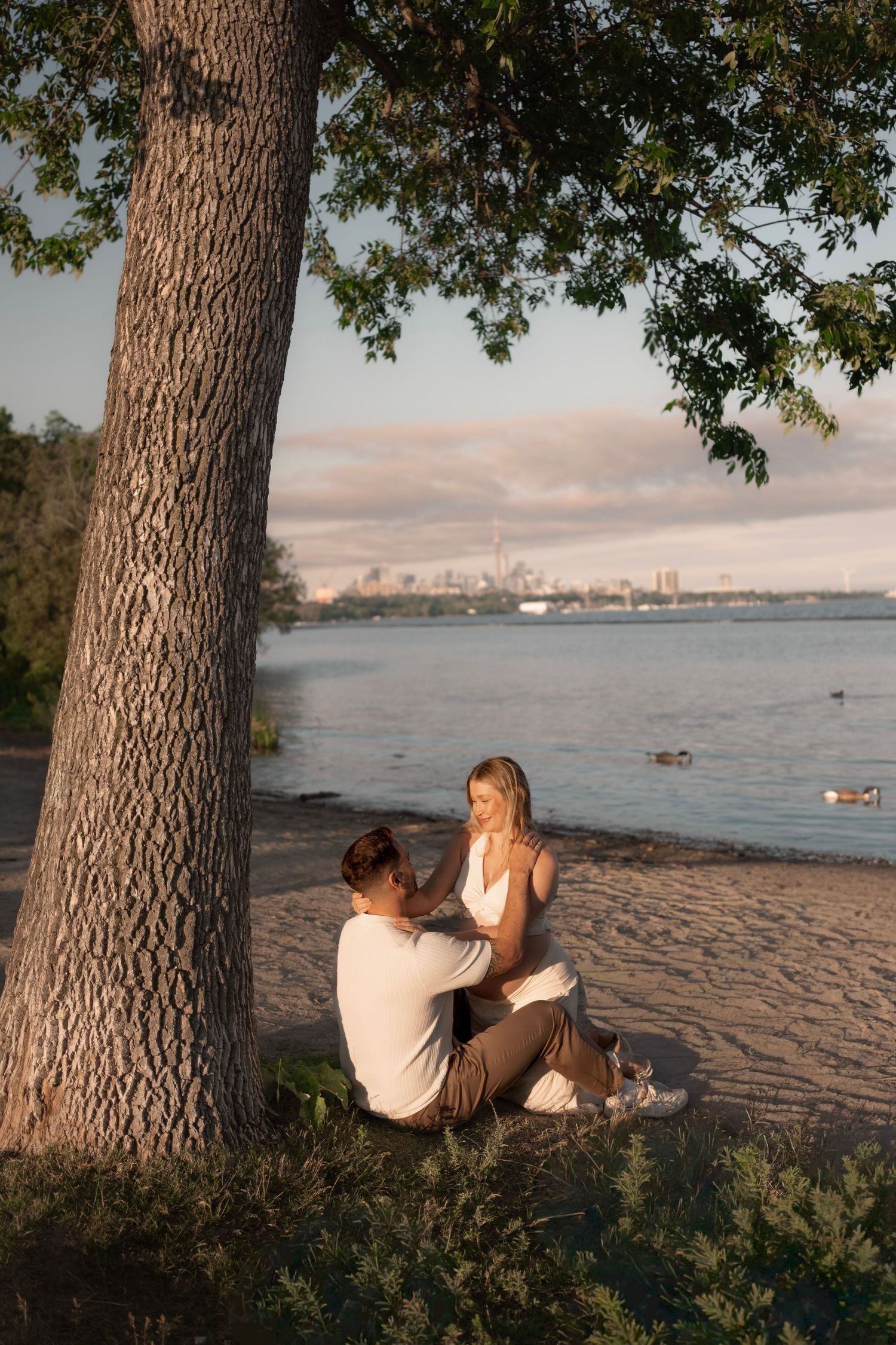 Couple sitting together under a large tree on a sandy beach at sunset with a city skyline in the background.