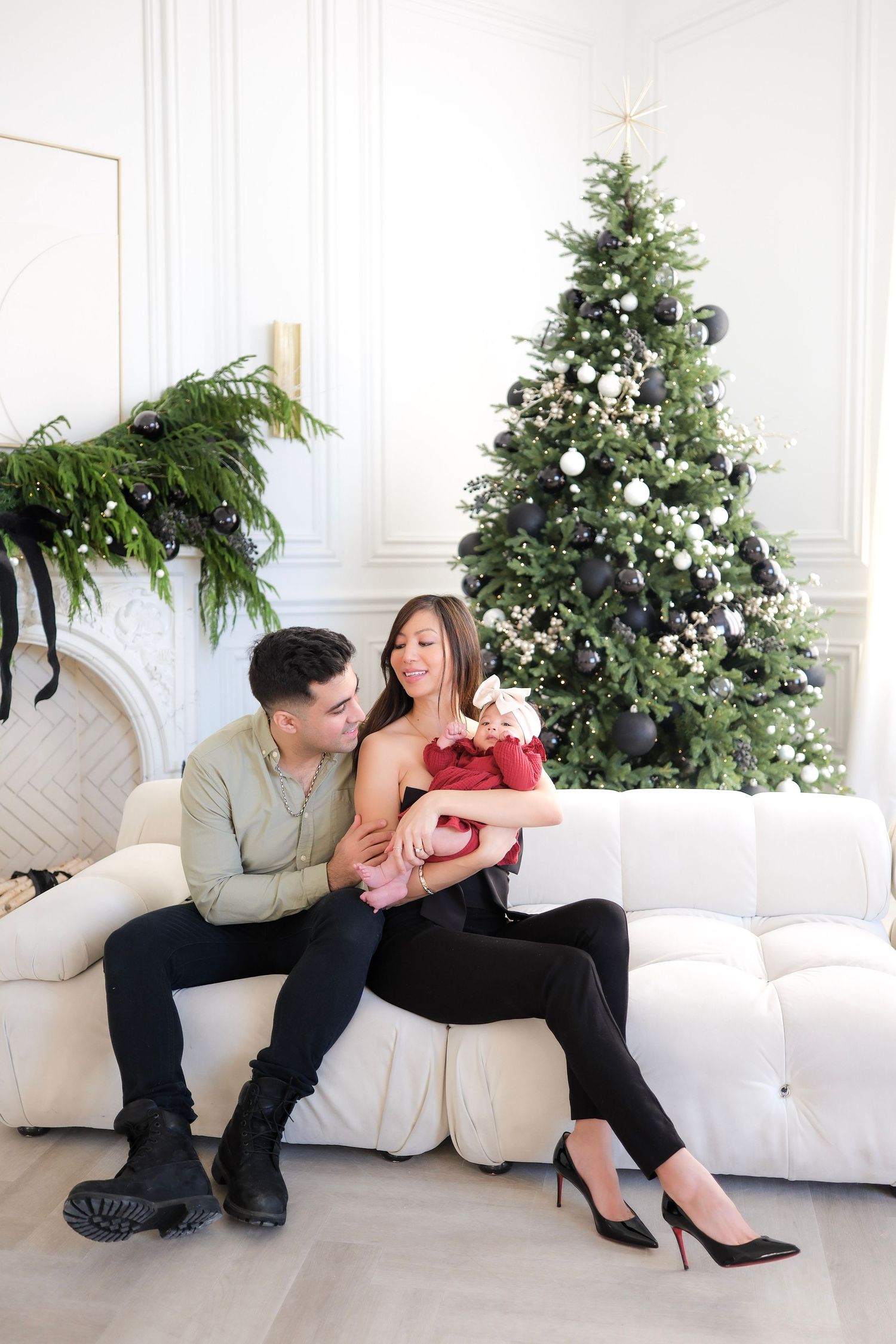 Couple in festive attire sharing romantic moments on white couch with decorated Christmas trees and greenery in background.