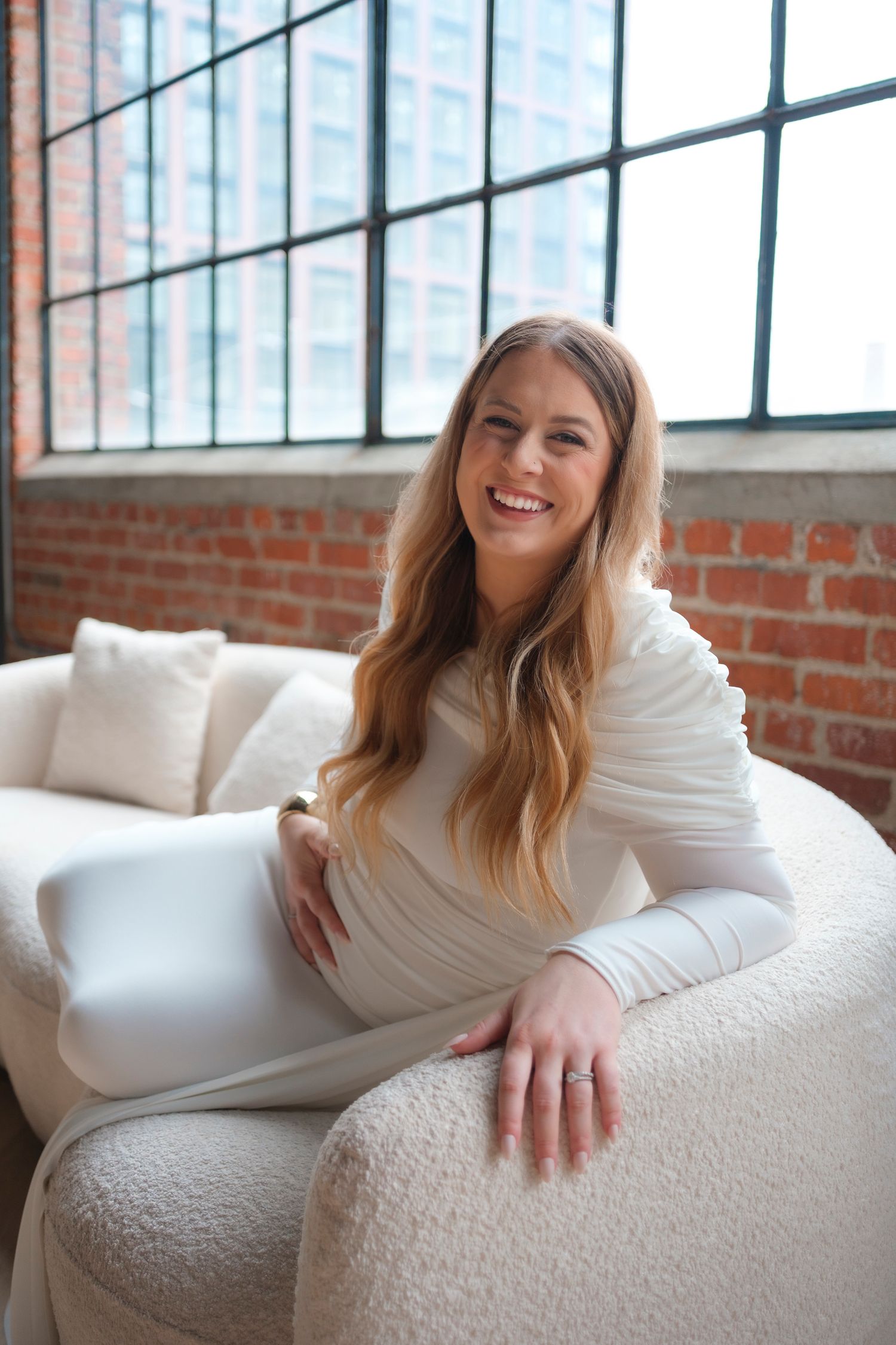 Woman in white long-sleeve dress sits smiling on plush white couch in industrial loft with exposed brick and windows.
