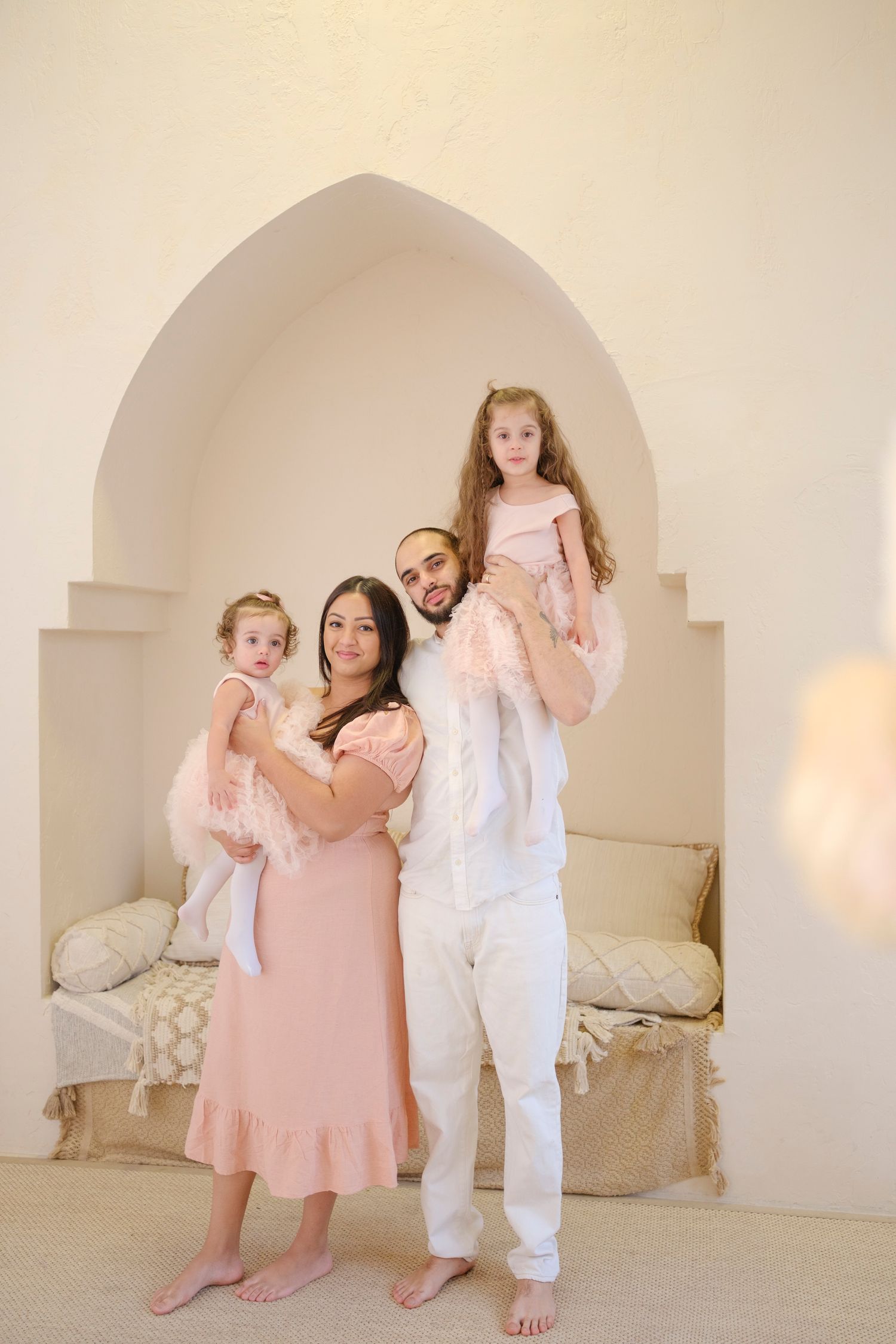 A family poses together in matching blush pink and white outfits inside an arched alcove with soft lighting.