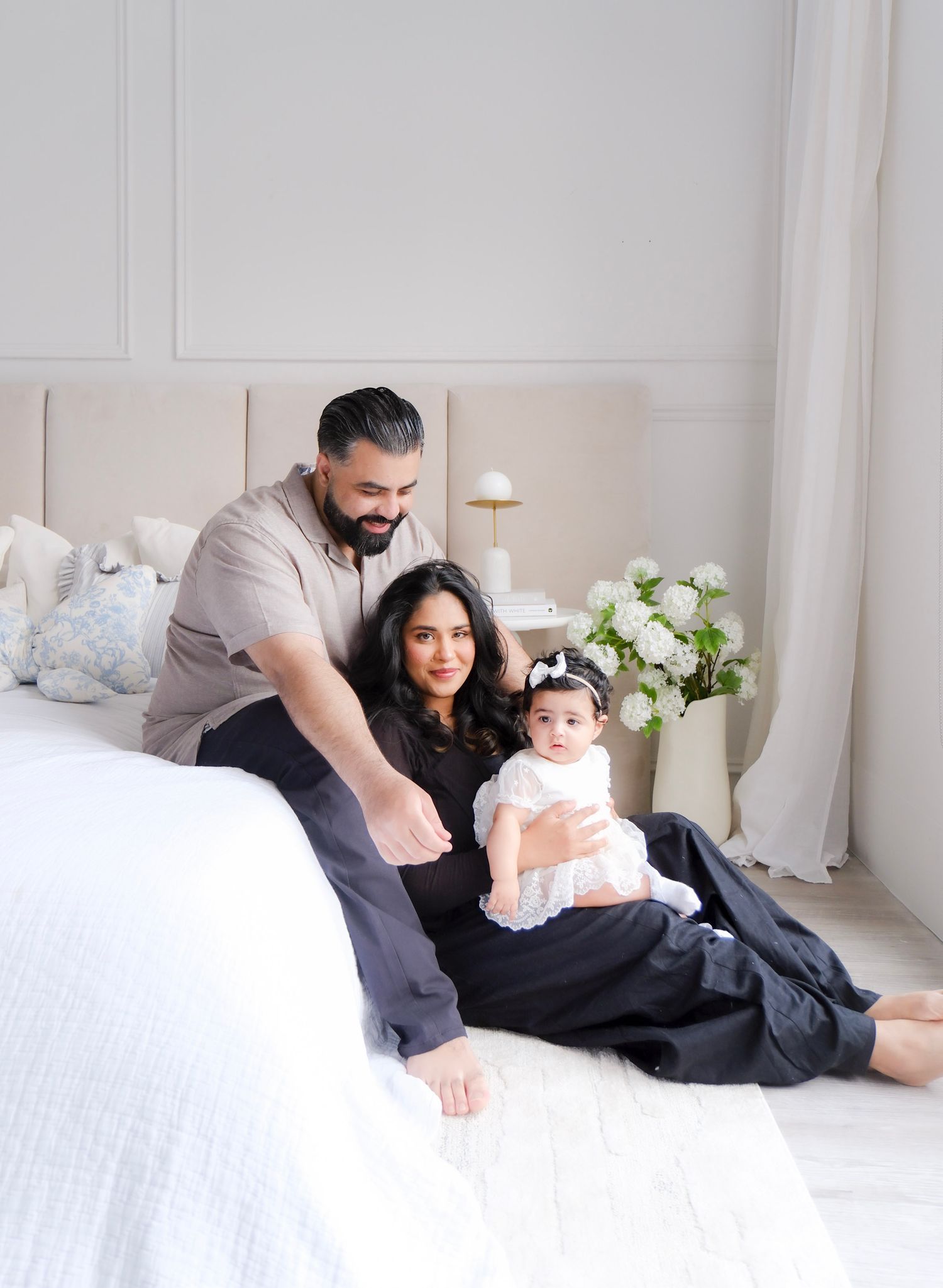 A family relaxes together on a white bed with decorative pillows and white flowers in a bright, modern bedroom setting.