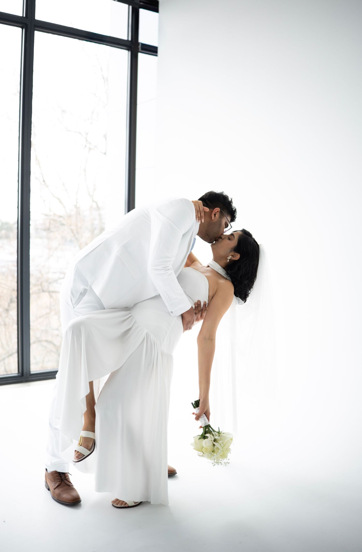 Groom in white suit dipping bride in white dress for a kiss near large studio windows.