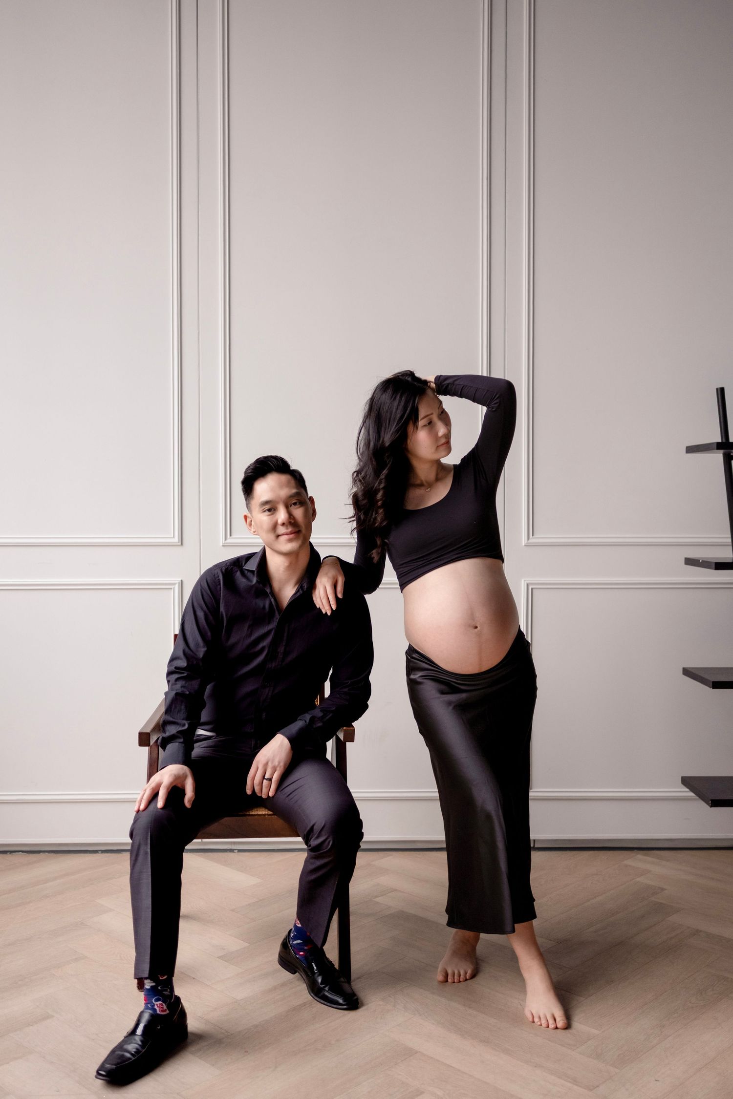 A pregnant woman in a black crop top and satin skirt poses with a seated man in a black shirt in an elegant studio.