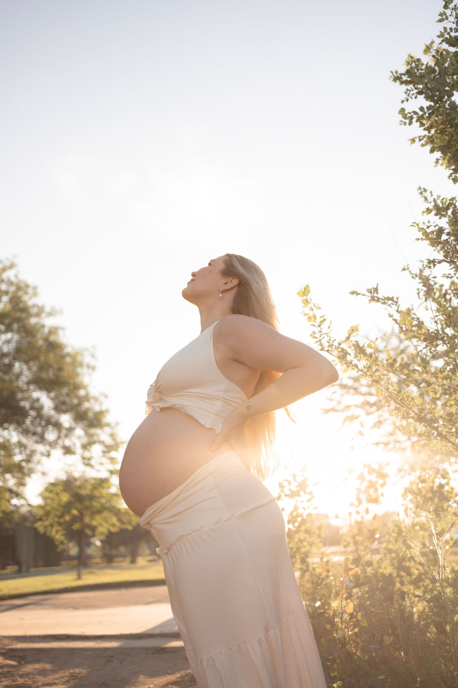 Pregnant woman in white dress holds her back while basking in golden sunset light outdoors.