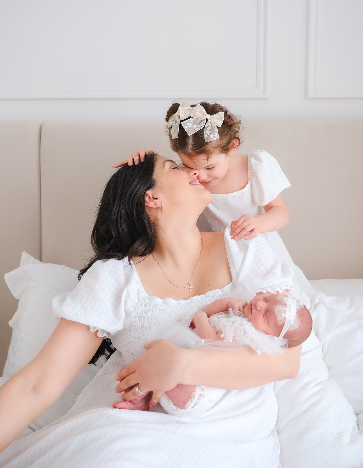 Tender family moment shows mother in white dress resting on bed with two children in coordinating white outfits.