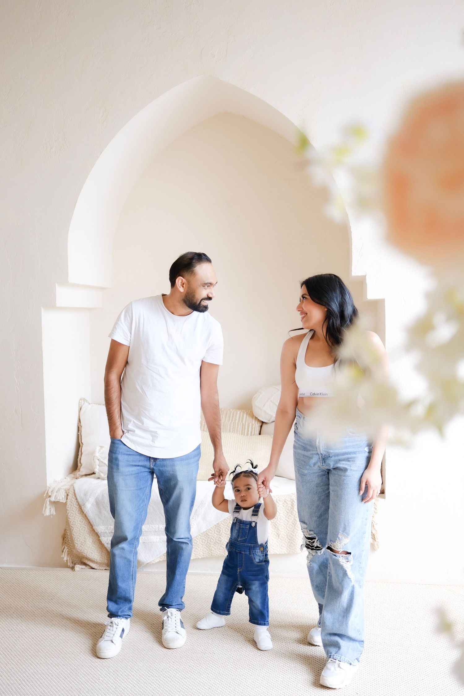A loving family walking together in a bright white interior space while holding their toddler's hands.