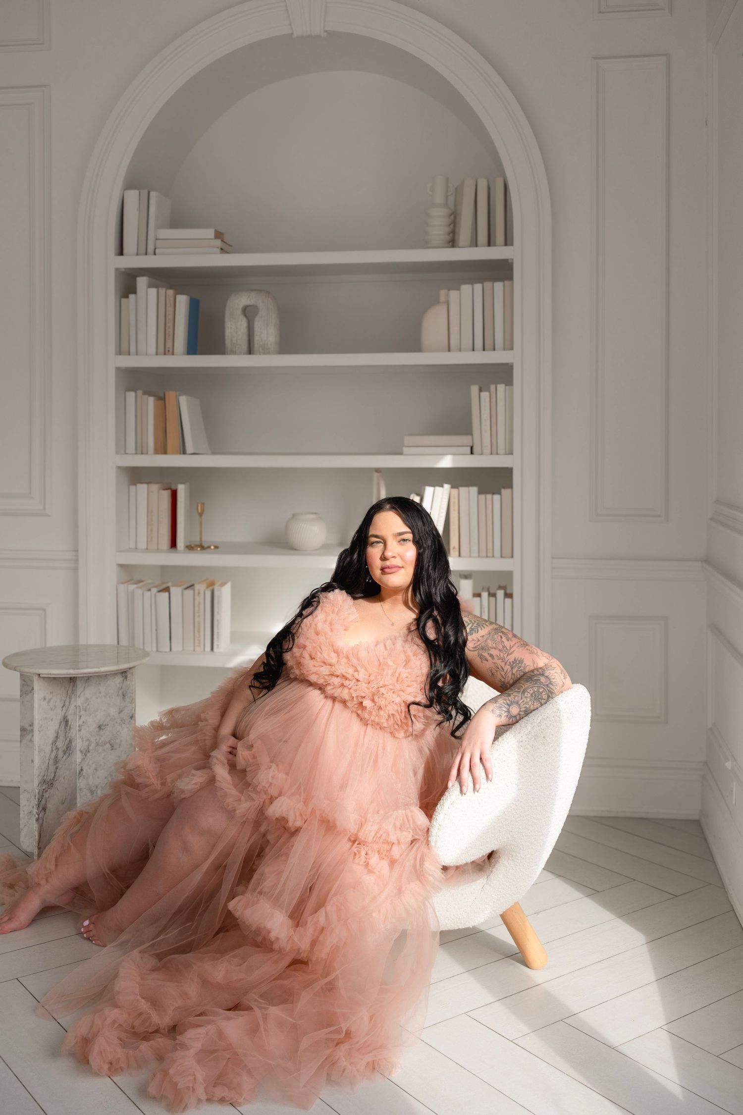 A woman in a flowing pink tulle gown sits gracefully in a white chair before an elegant arched bookshelf.