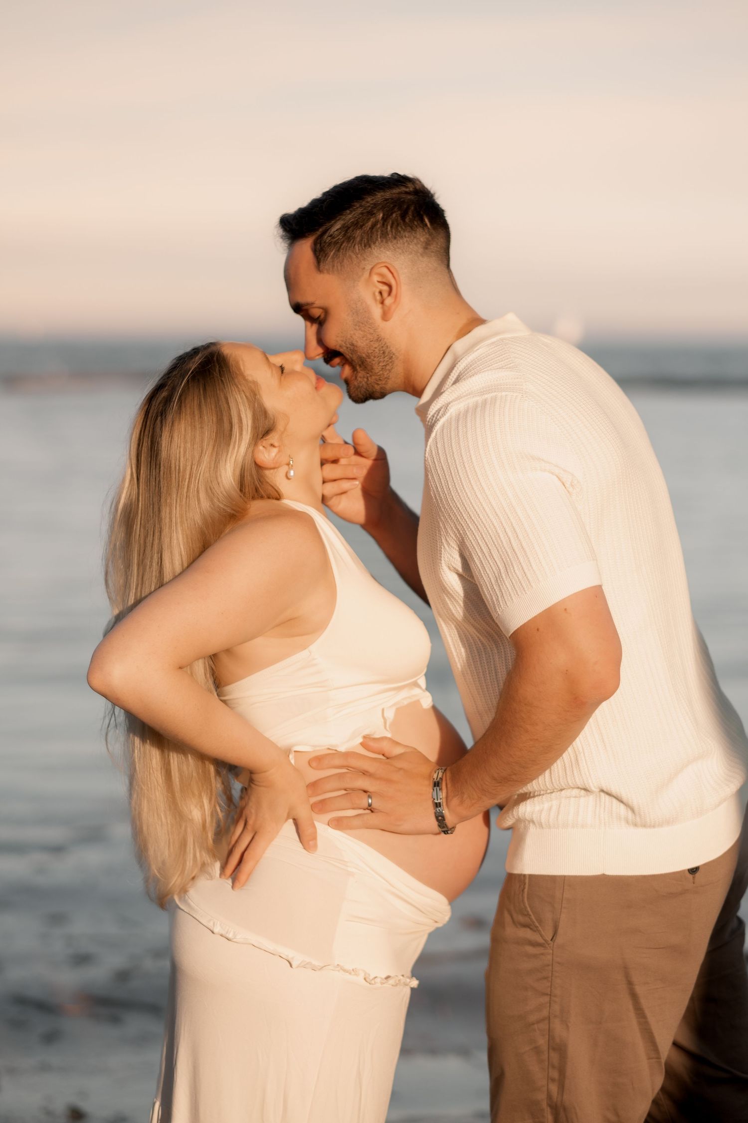 Couple sharing a romantic moment at beach sunset, man kissing pregnant woman's forehead while holding her bump.
