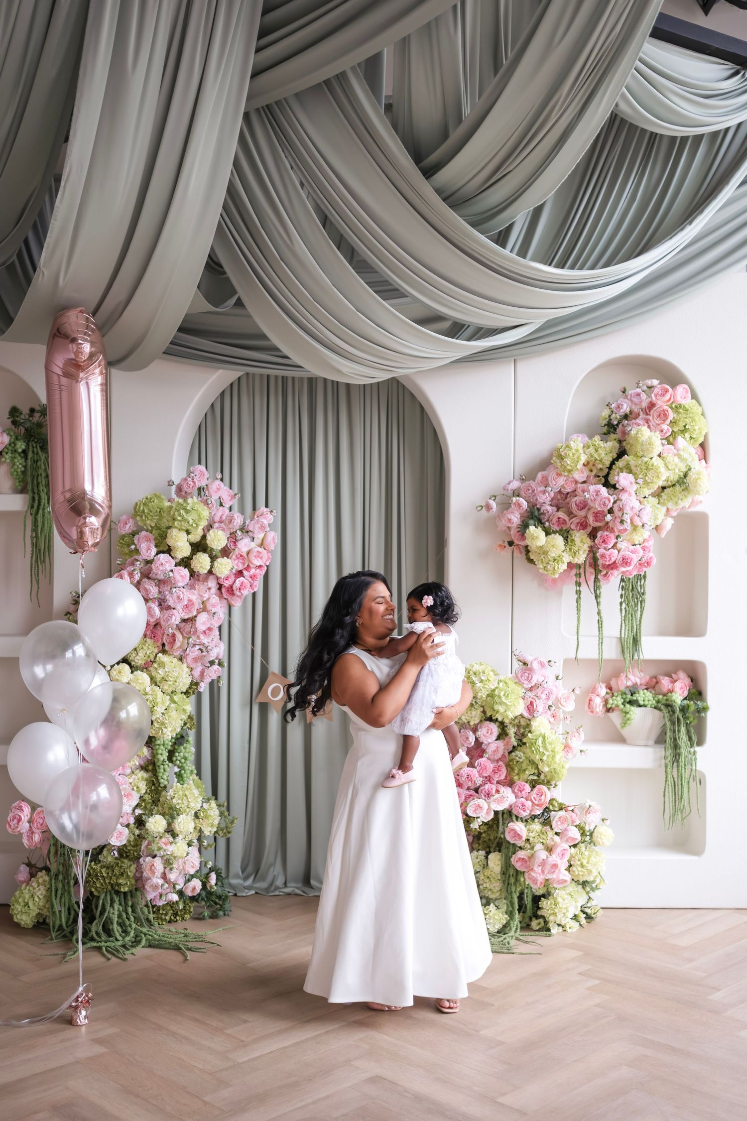 A mother in a white gown holds her baby at a floral first birthday celebration with pink blooms and balloons.
