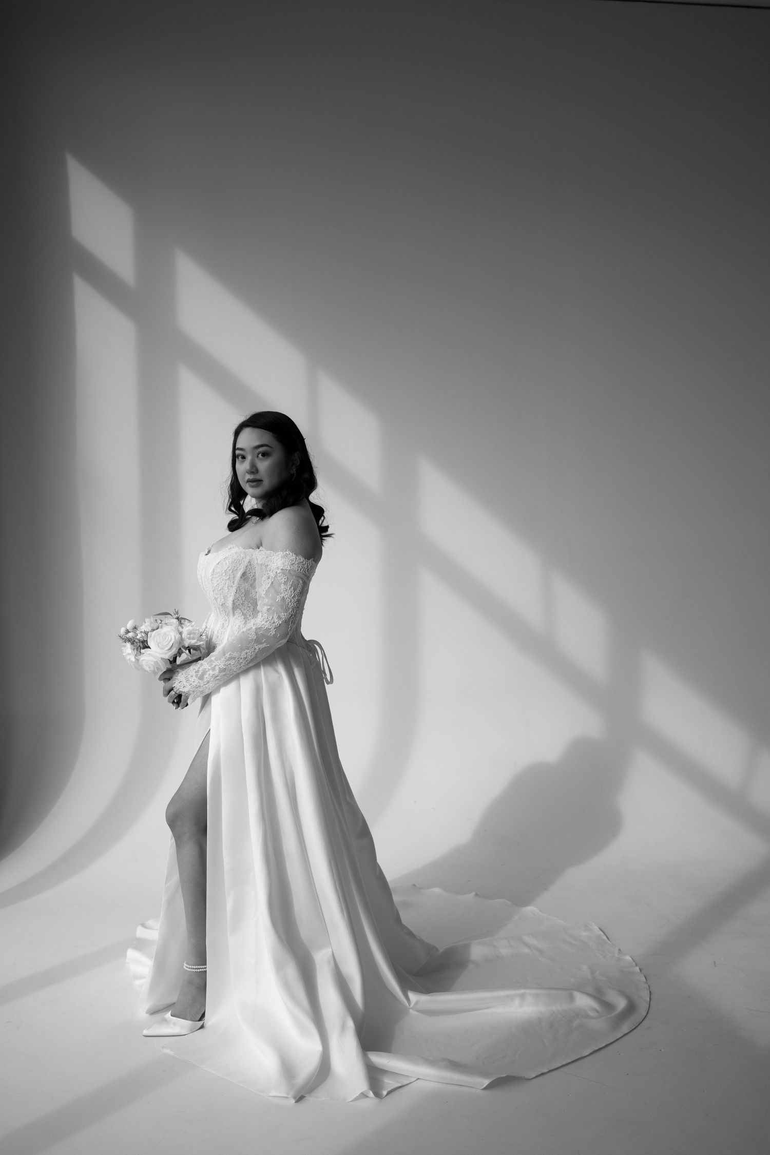 Elegant bride in off-shoulder lace gown with train holds bouquet in dramatic black and white light.