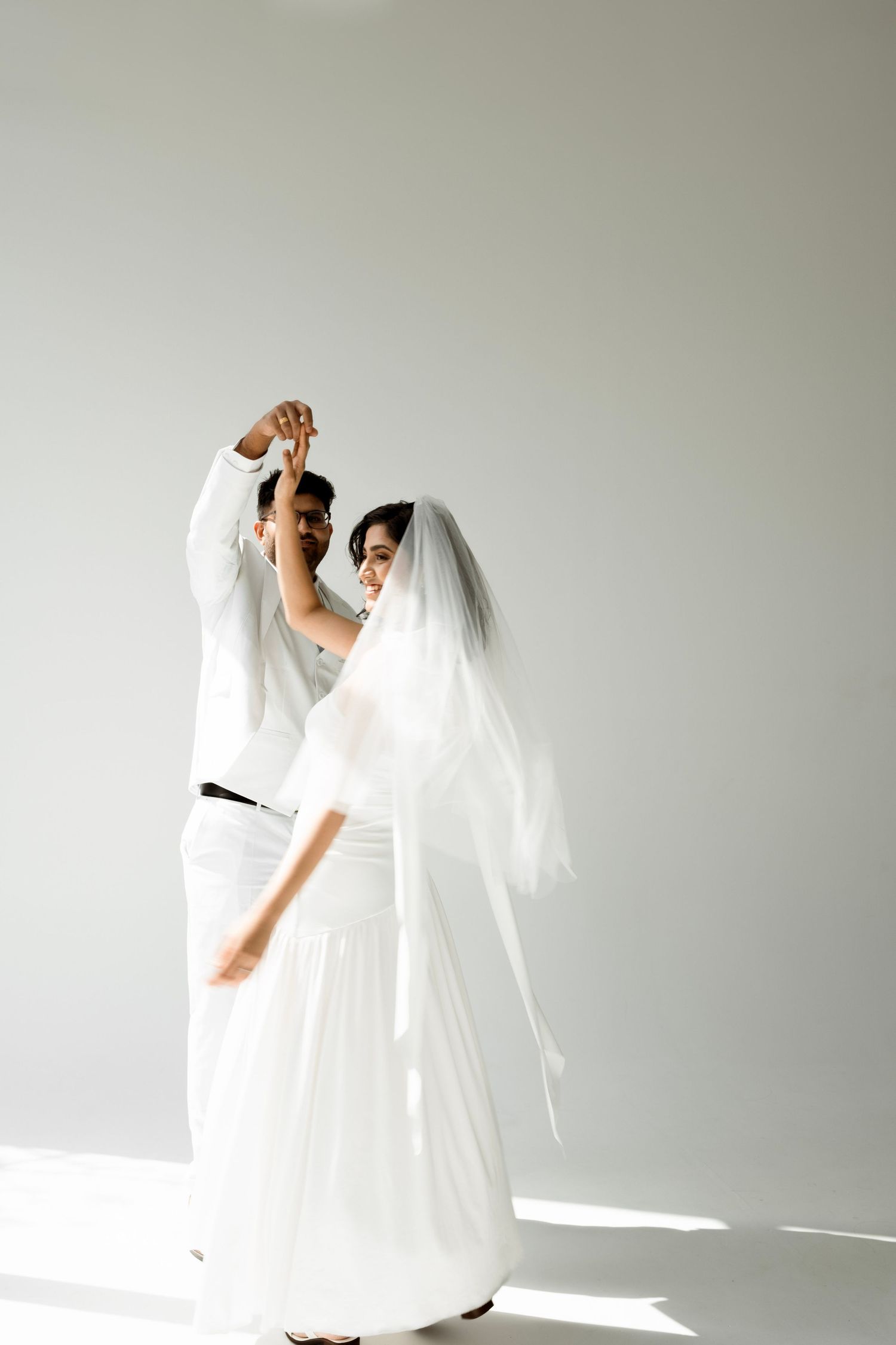 Bride and groom dancing together in matching white outfits, bride wearing a flowing veil, against a white background.