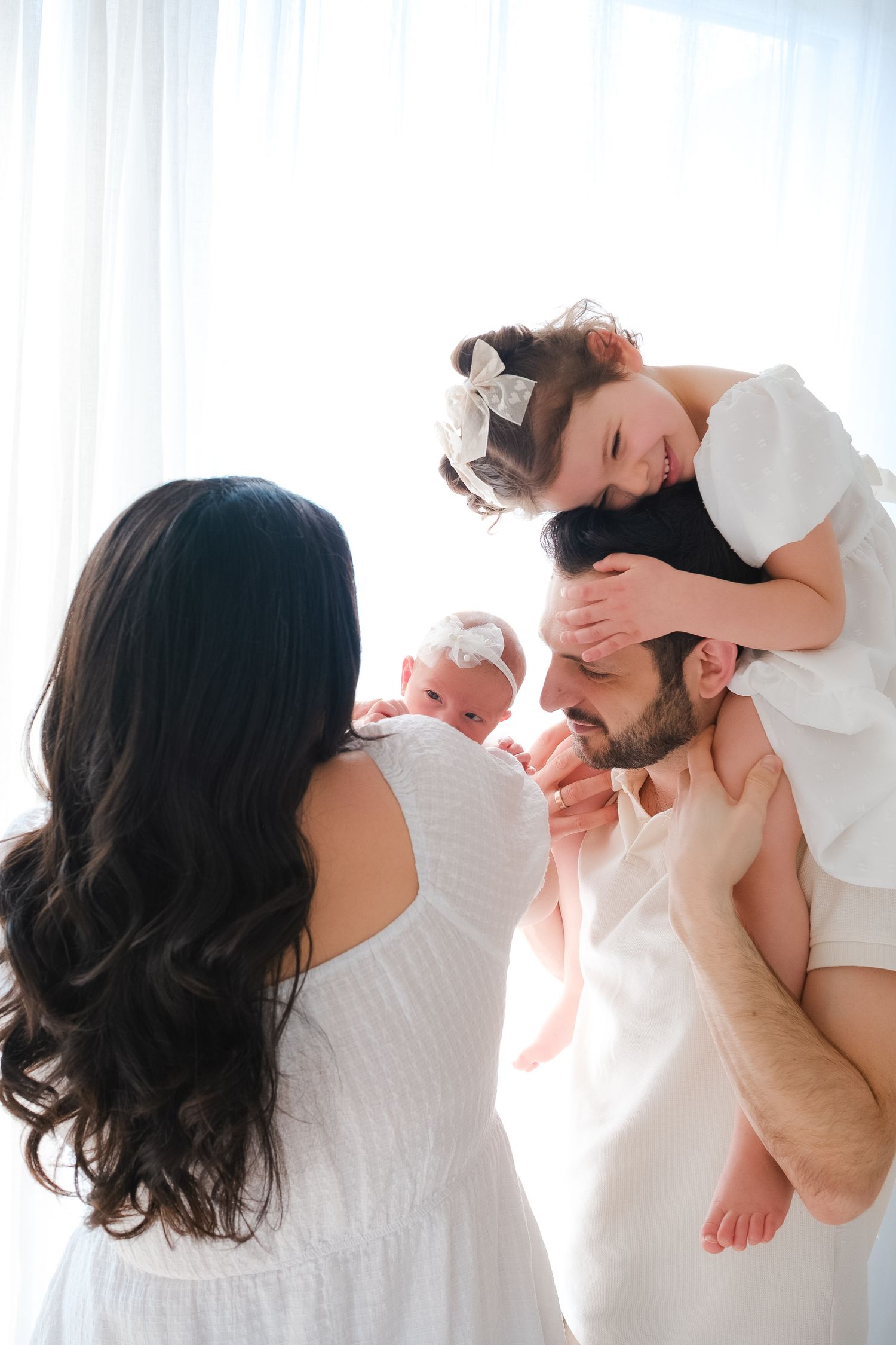 A family wearing white clothes embraces and shares a joyful moment together by a bright window.