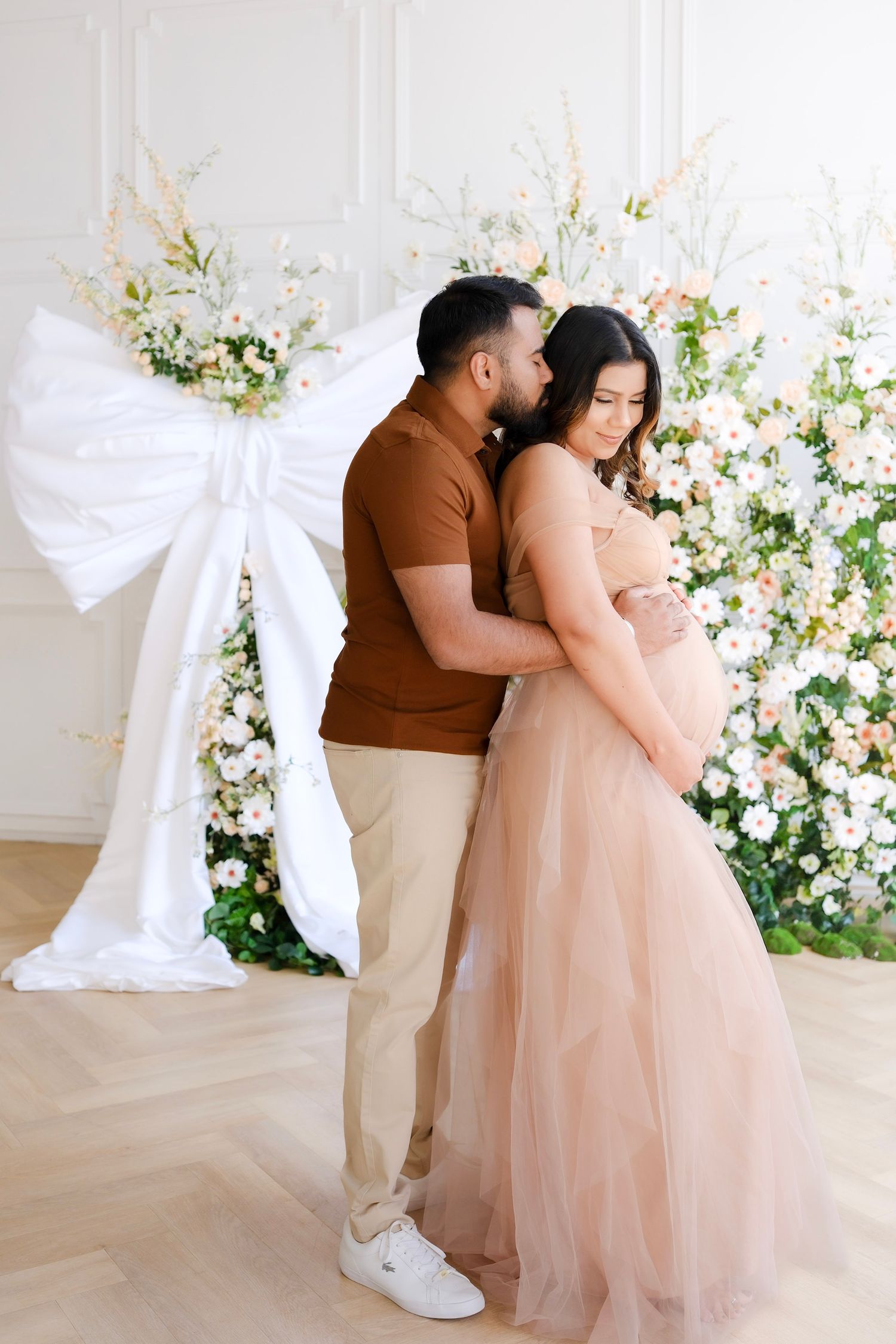 A couple poses against a floral backdrop with white bow decorations while wearing brown and blush formal attire.