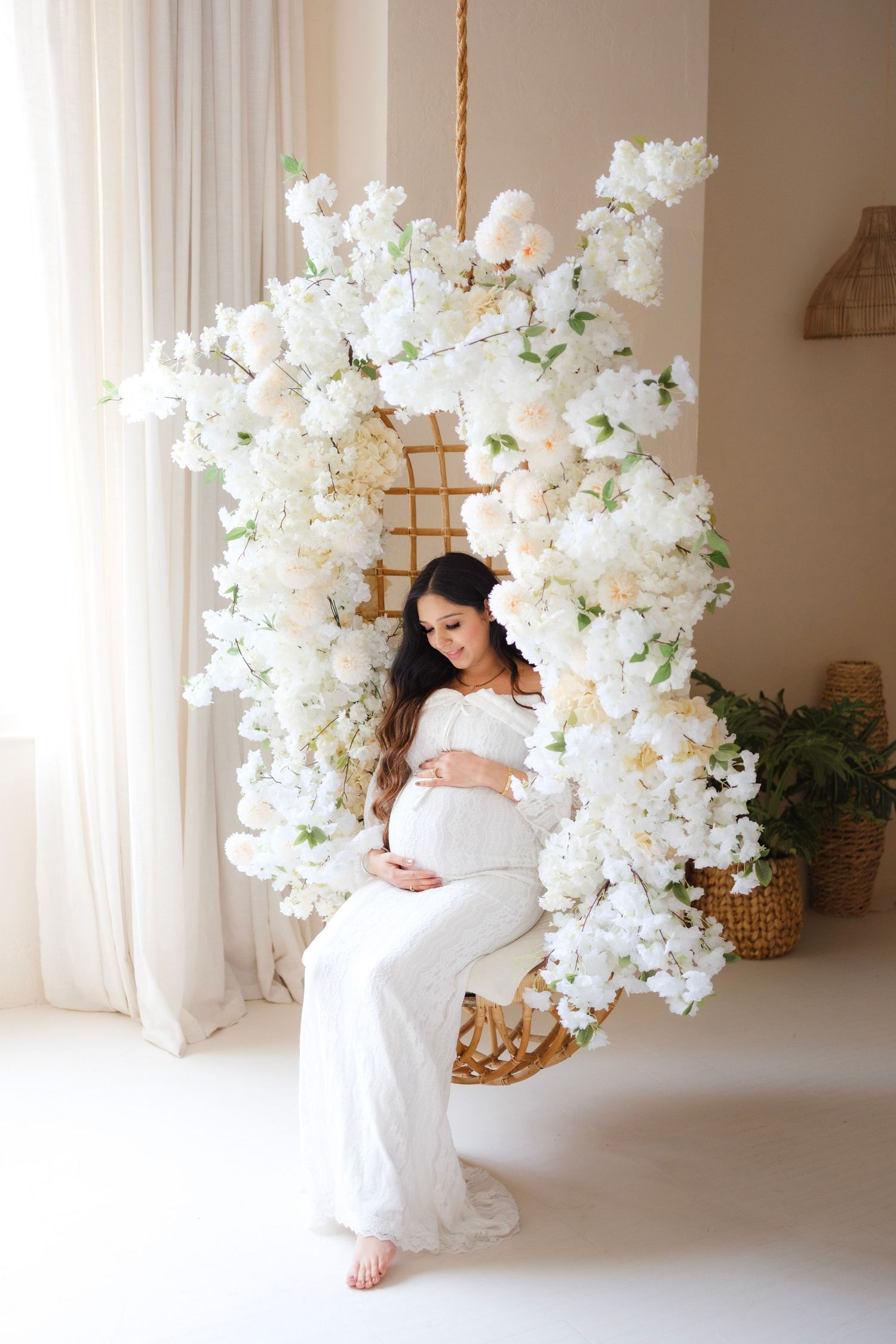 Pregnant woman in white dress sitting on floral swing chair surrounded by white blooms in bright studio.