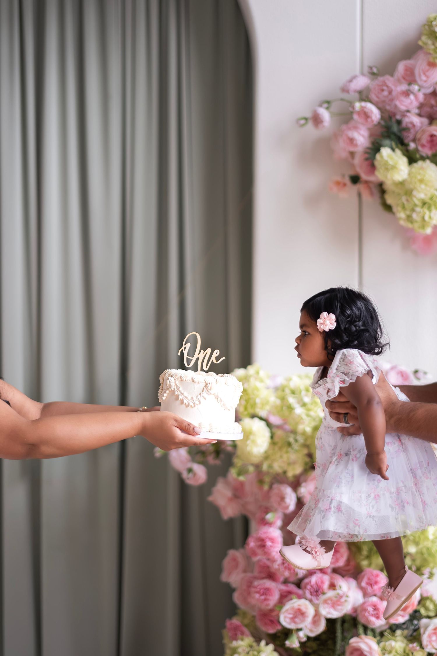 A baby girl in a white dress reaches for her first birthday cake with a gold ‘One’ topper amid pink flowers.