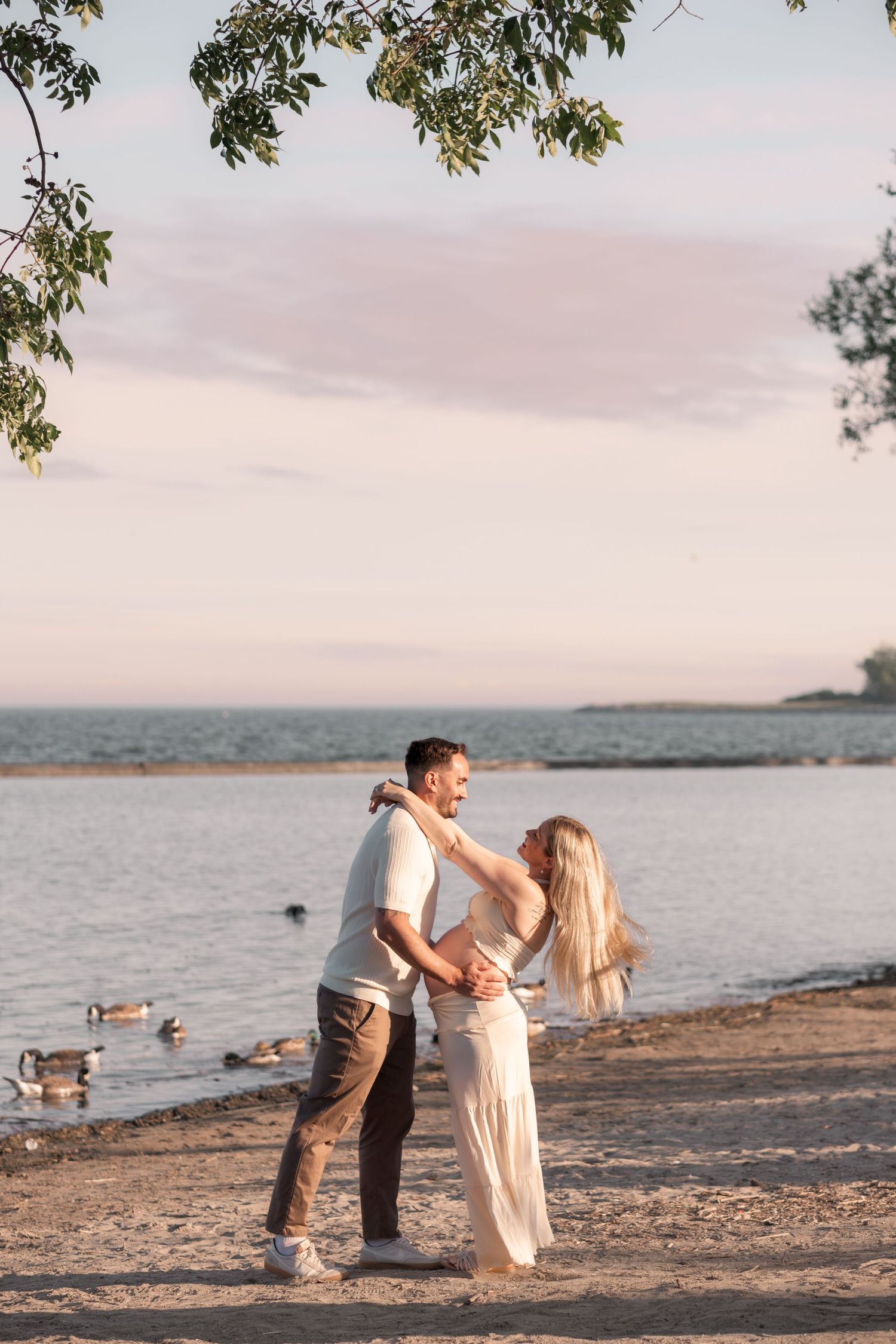 A couple embraces romantically on a sandy beach at sunset with calm water and geese in the background.