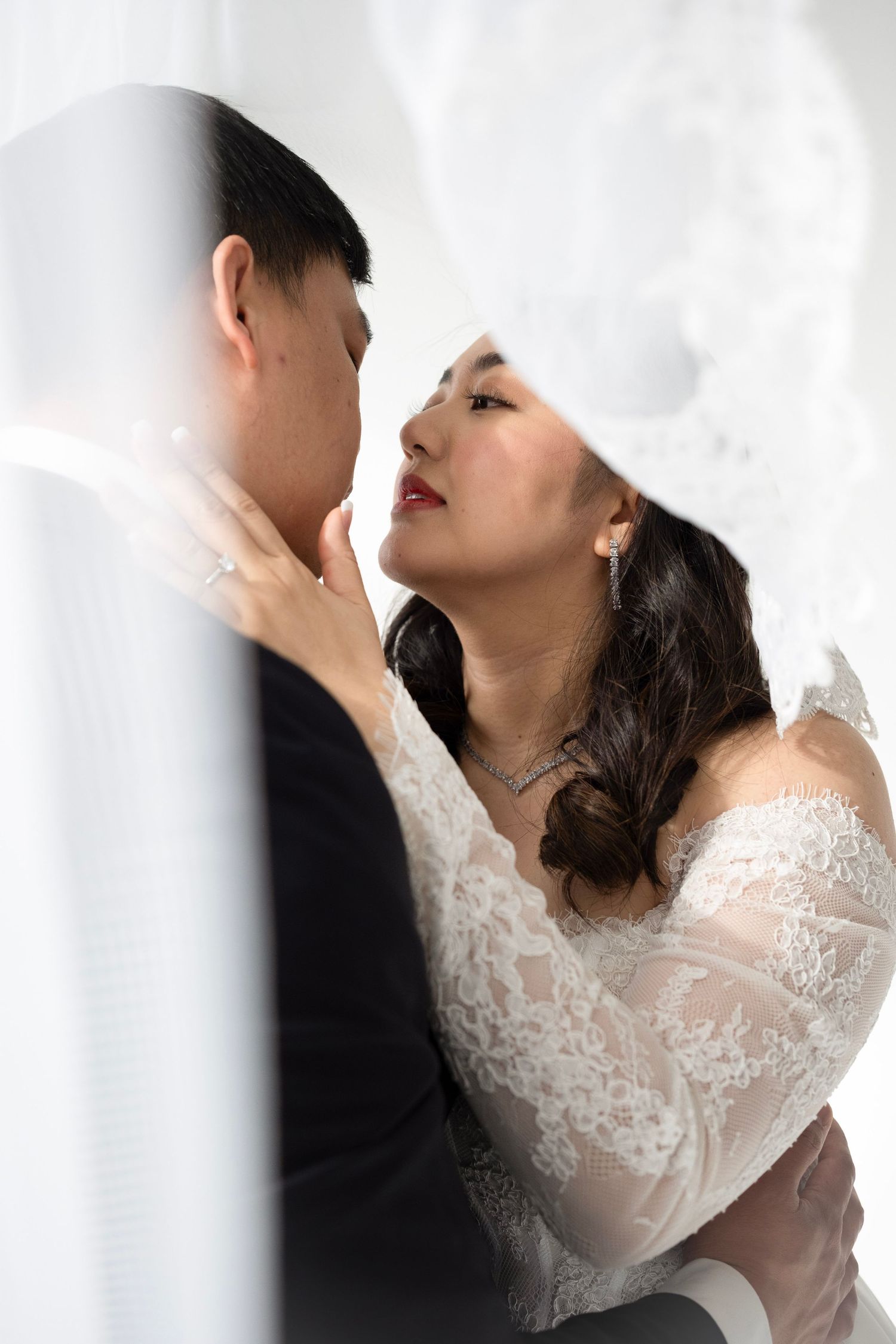 Bride in lace off-shoulder wedding dress gazes lovingly at groom under veil in an intimate close-up portrait.