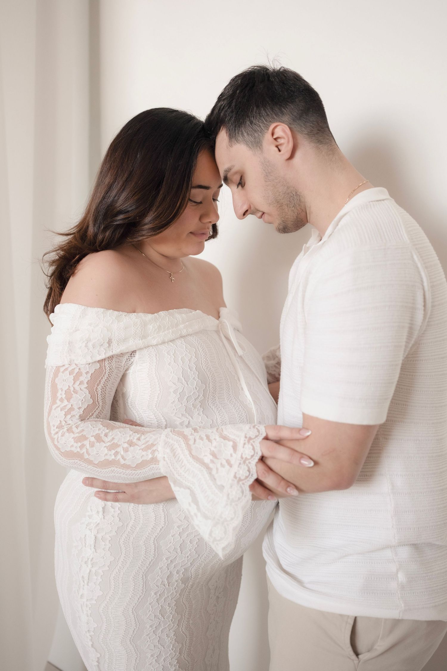 Pregnant woman in white lace off-shoulder gown and man in white shirt tenderly touching foreheads in soft studio light.