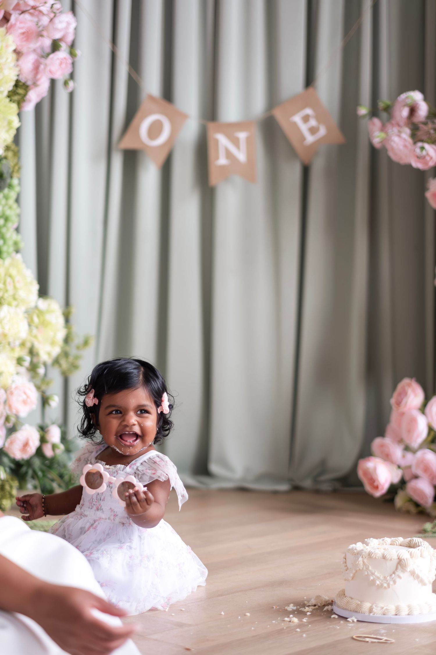 A baby girl in a white lace dress celebrates her first birthday with a smash cake and floral decorations.