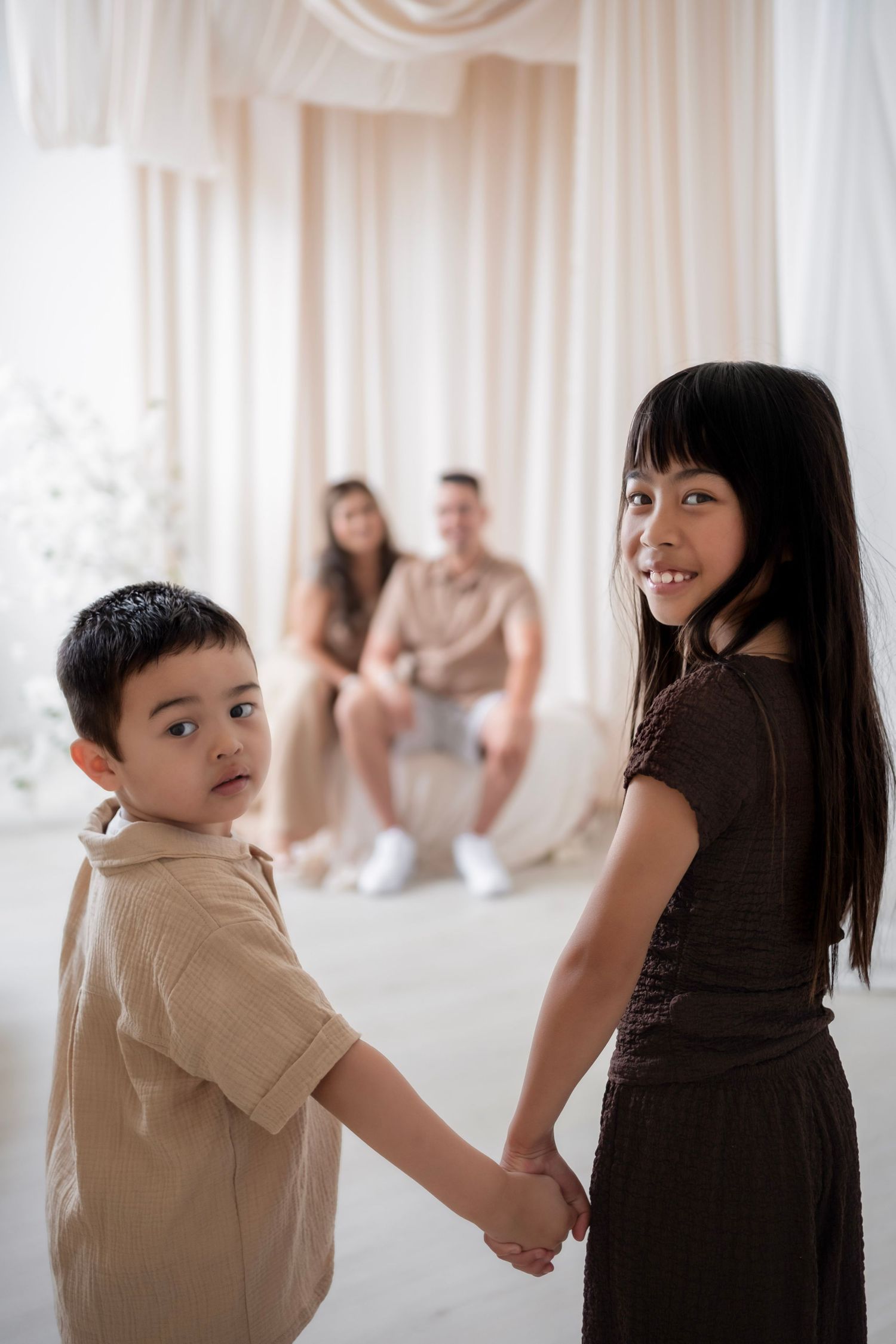 Two Asian children holding hands and smiling in front of their parents in a bright white studio setting.