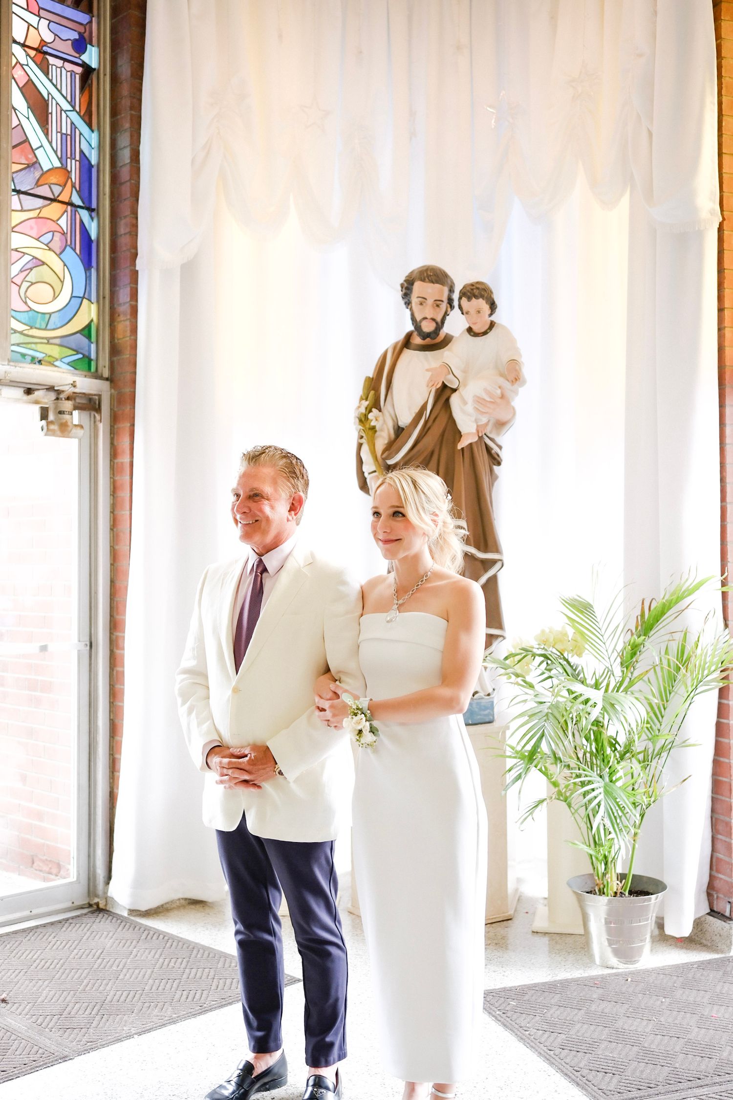 A couple in a church stands before a religious statue during their wedding ceremony, with stained glass windows visible.