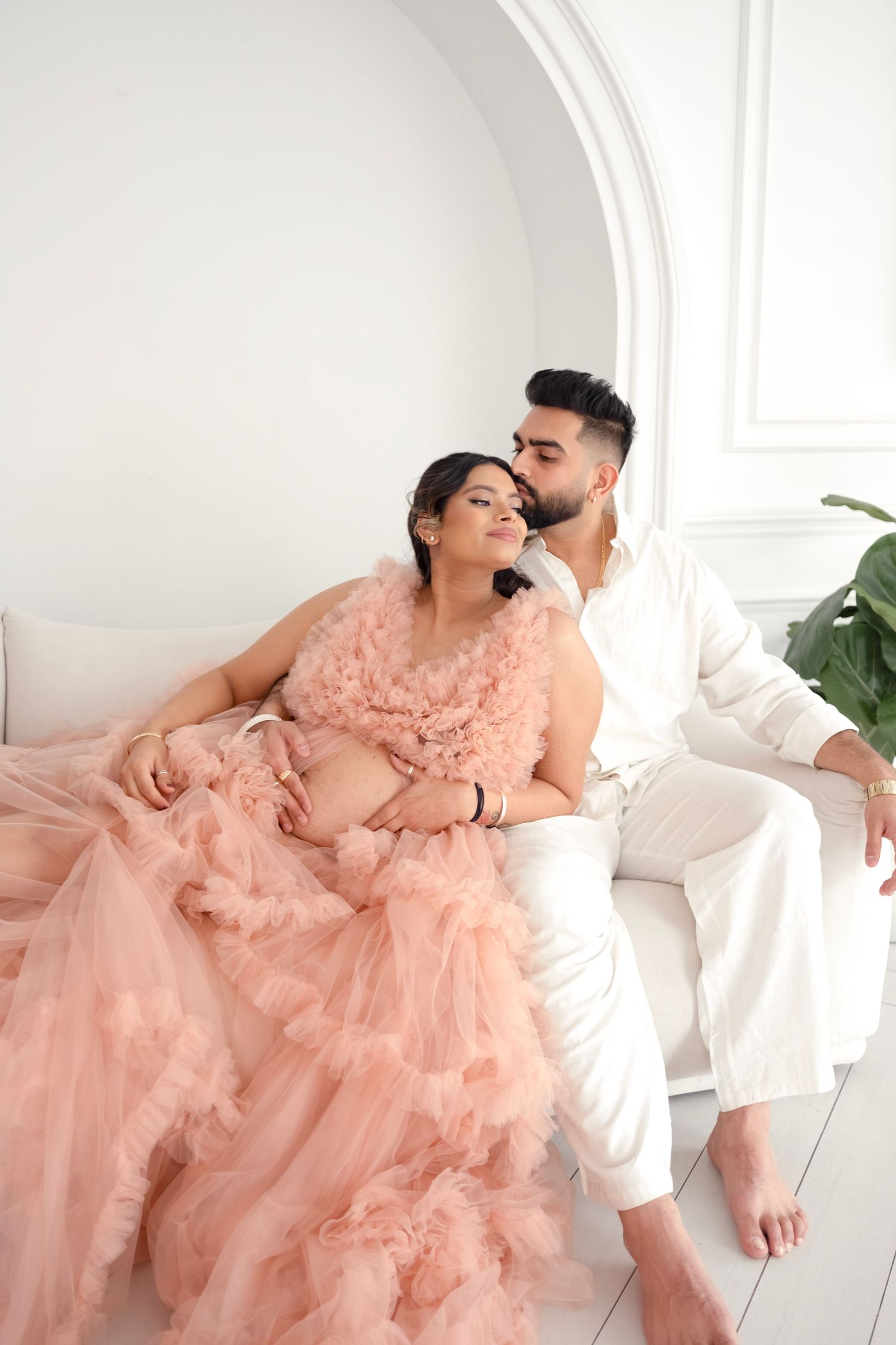 A couple in a romantic maternity photo shoot with a flowing pink tulle gown against a white arched backdrop.