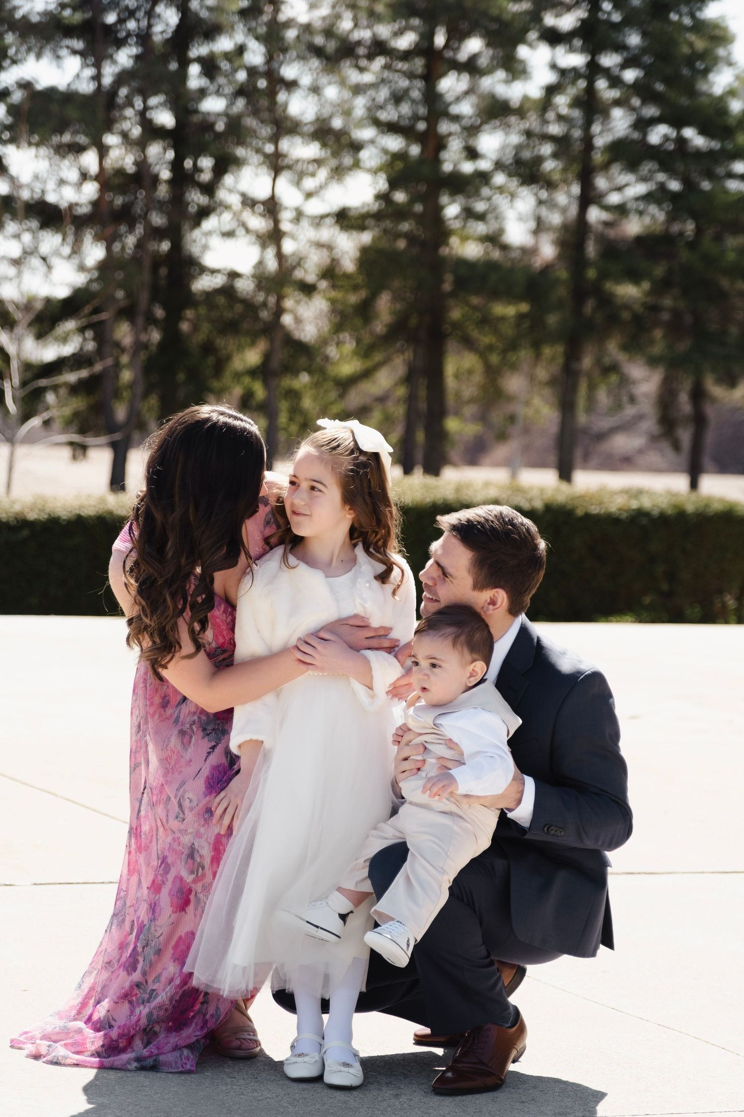 A family of four poses outdoors, parents crouching with a girl in white dress and baby boy, surrounded by tall pine trees.
