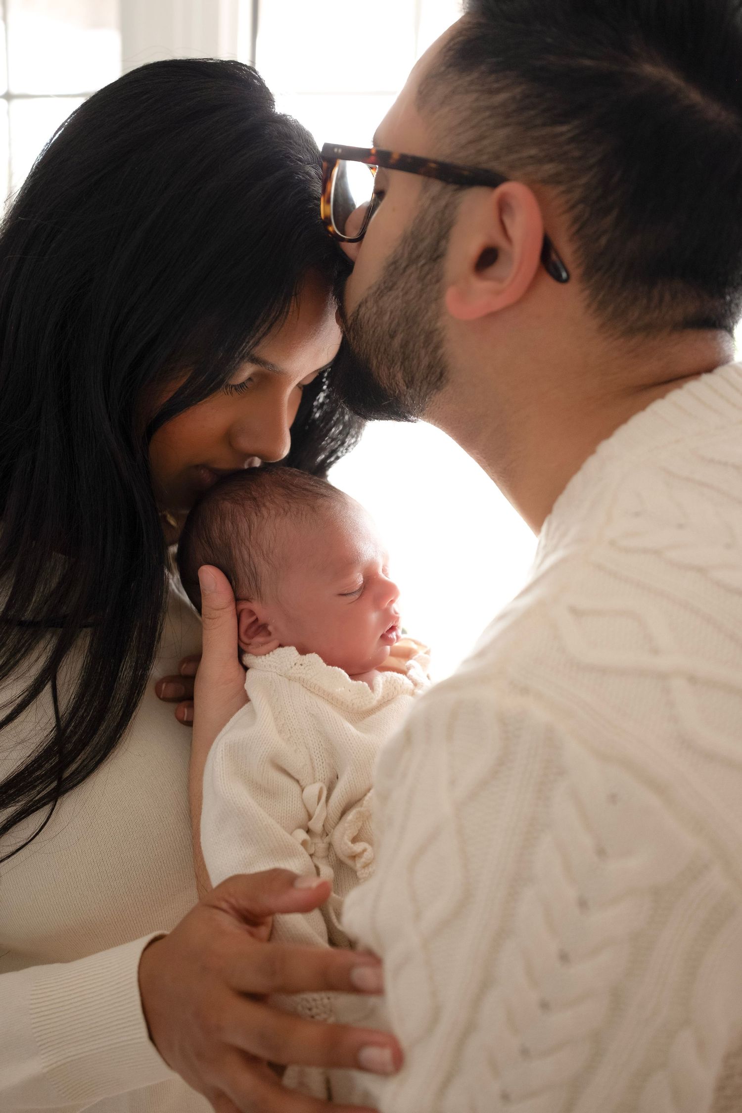 New parents tenderly kiss their sleeping newborn baby wrapped in white, sharing a precious family moment.