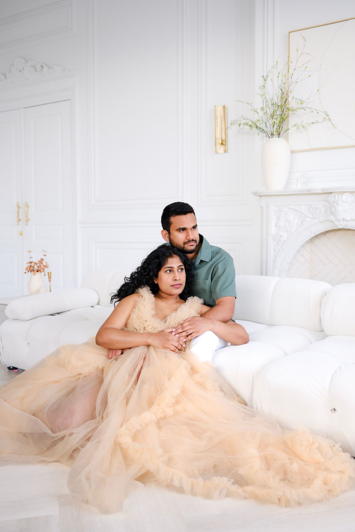 A couple poses together in an elegant white room, with the woman wearing a flowing champagne tulle gown.