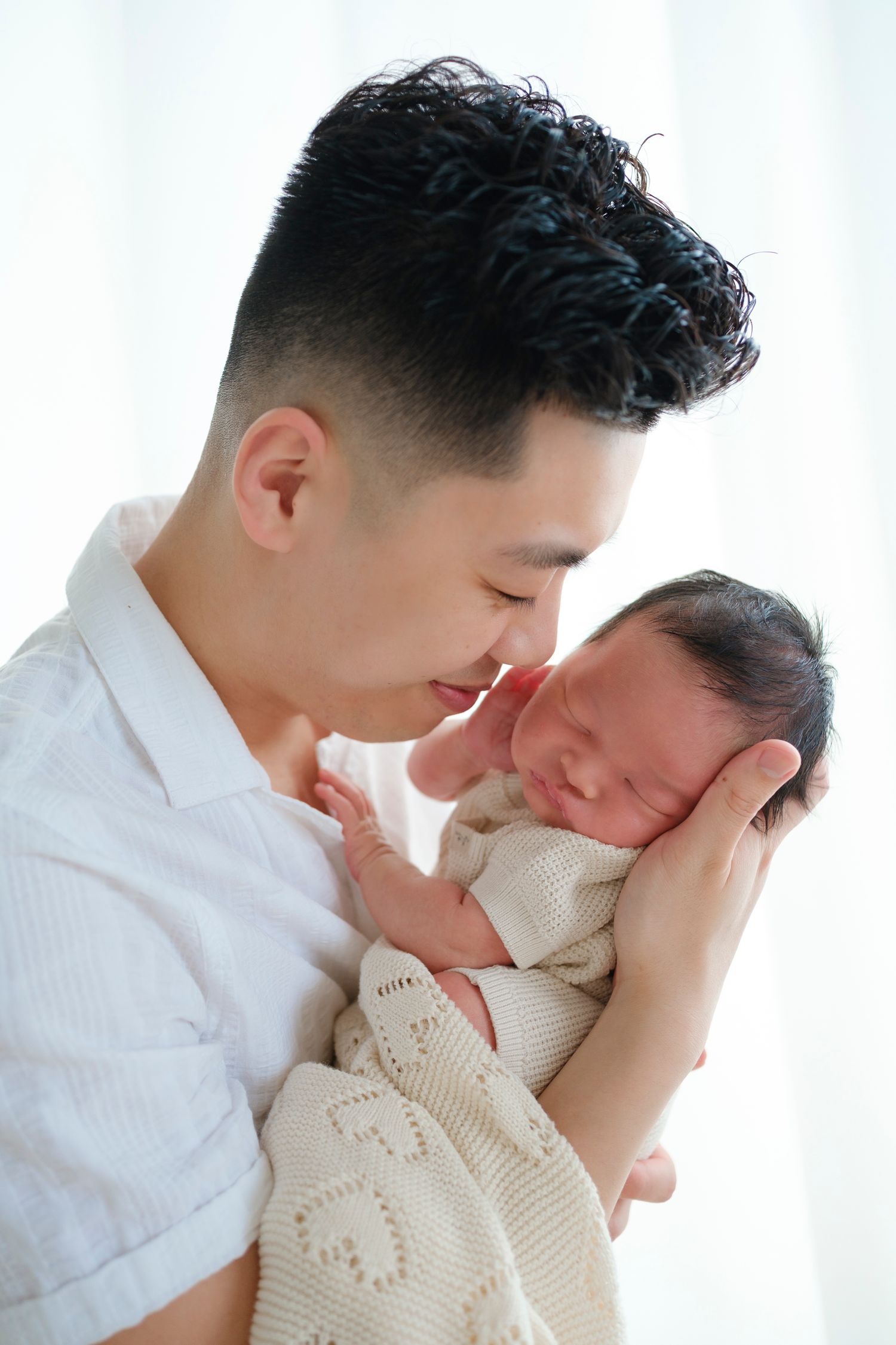 A parent tenderly holds and gazes at their sleeping newborn wrapped in a cozy knit blanket against a light background.