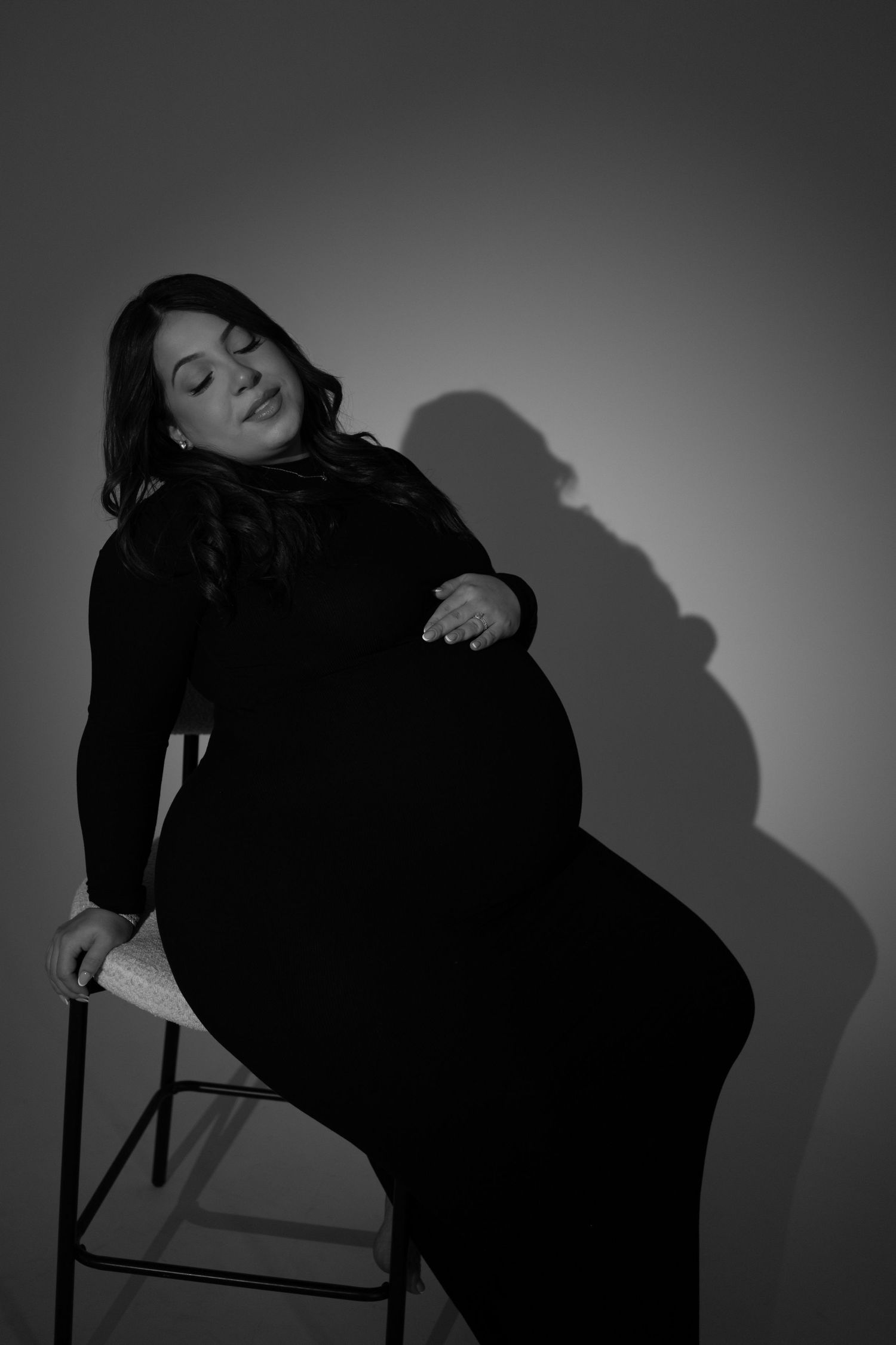 A black and white maternity portrait shows a pregnant woman in a dark dress sitting on a stool with dramatic shadow.