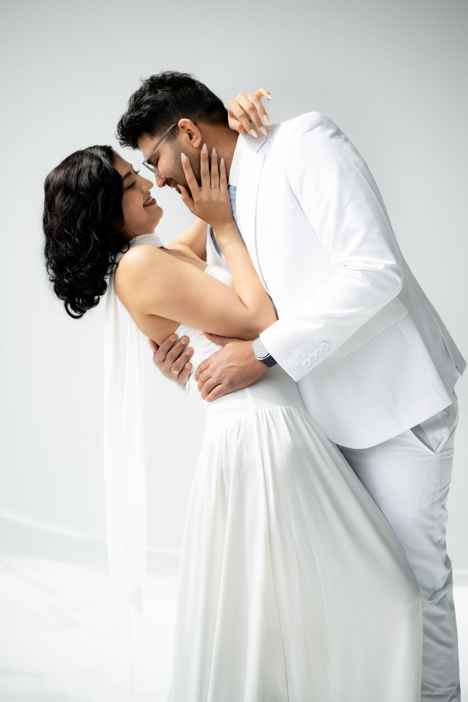 Bride and groom in white attire sharing a romantic almost-kiss embrace in a bright studio setting.