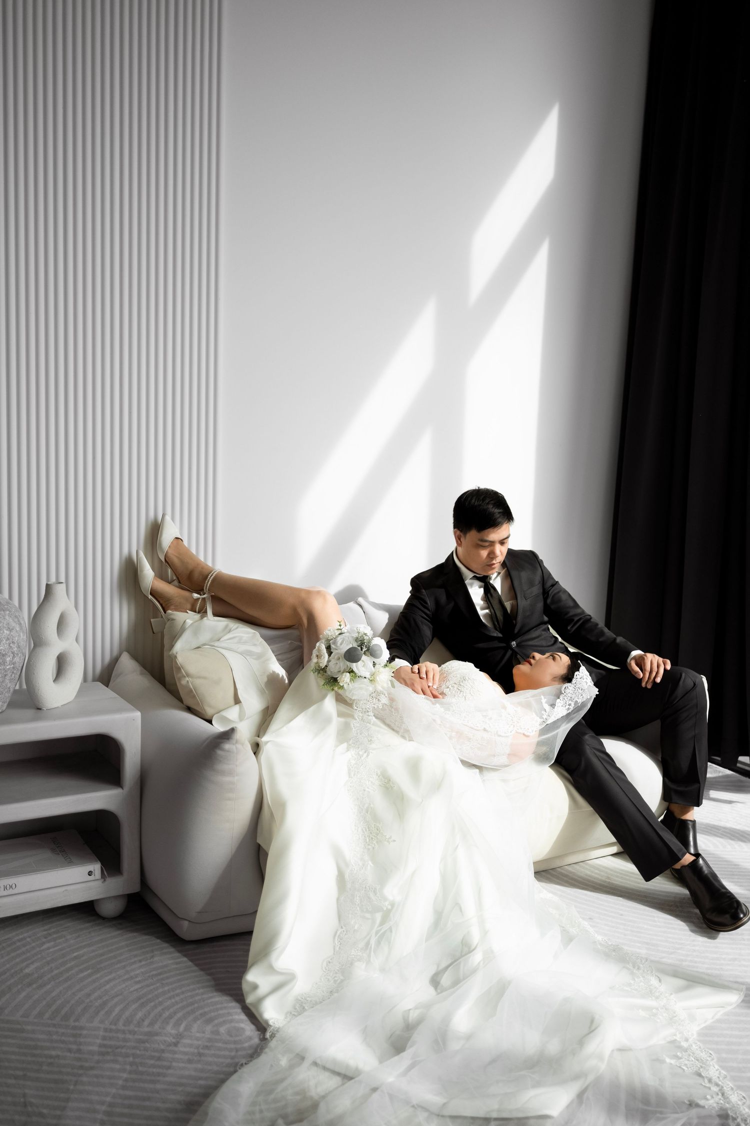 Bride and groom relaxing on a white sofa in elegant wedding attire, sharing a romantic moment in a modern bright room.