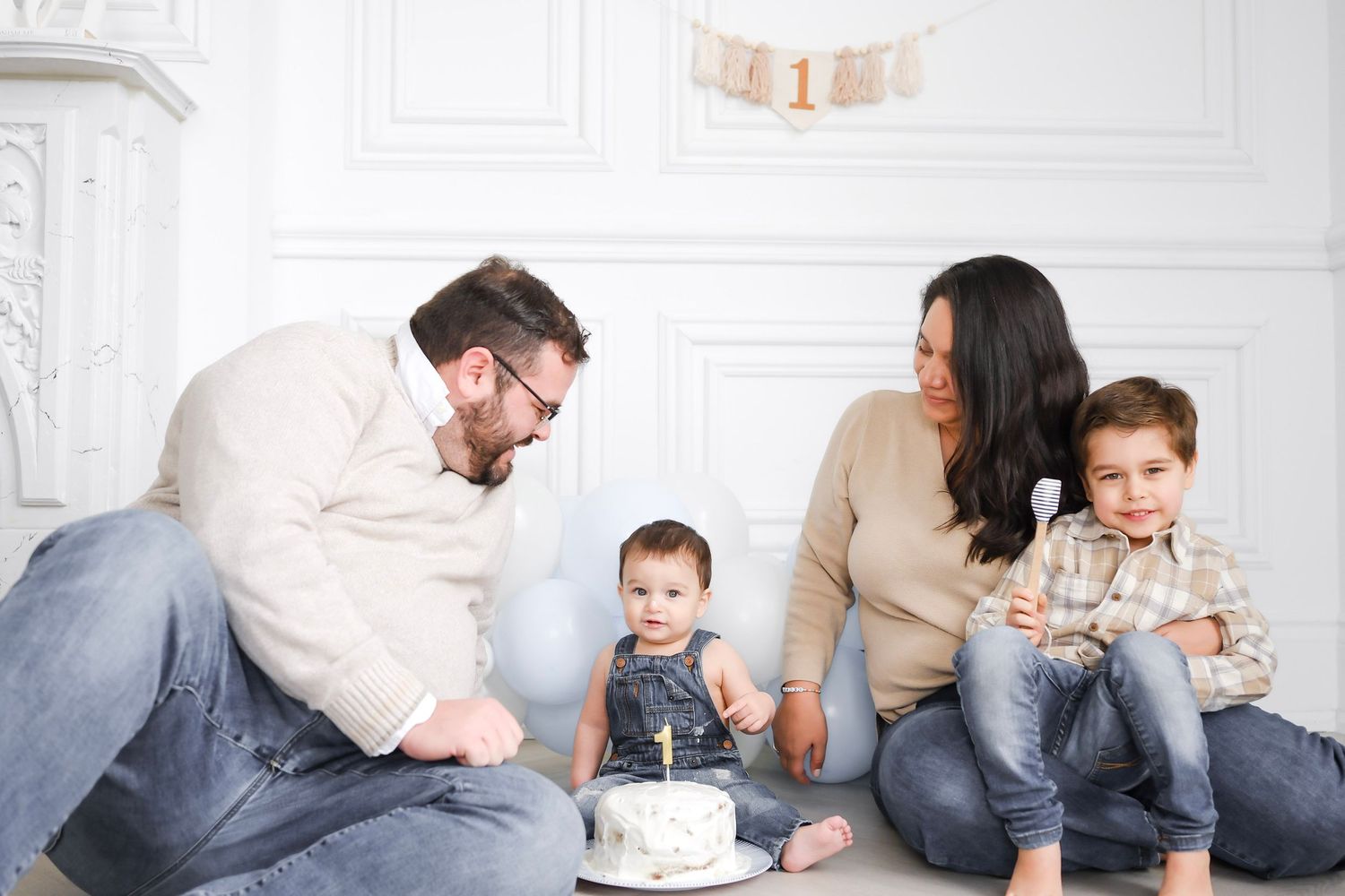 Family celebrating baby's first birthday with a small cake in a bright white room.