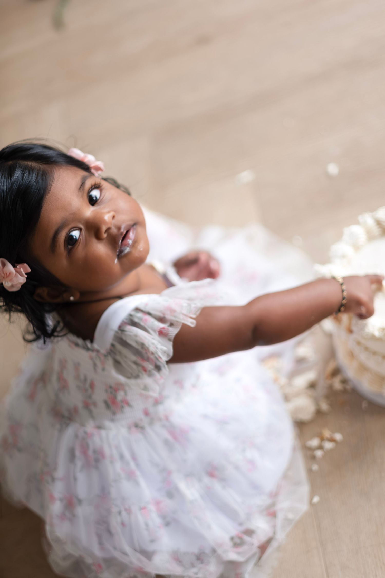 Young girl in white floral dress reaching toward a cake, looking up at camera during birthday celebration.
