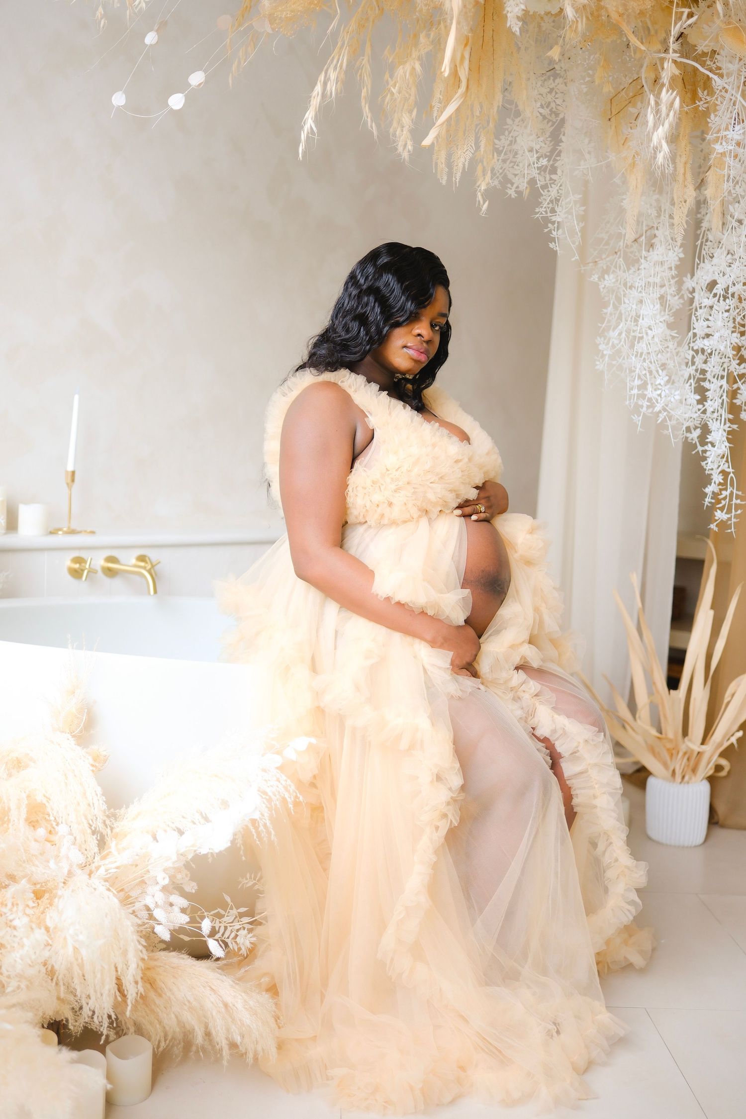 A maternity portrait in a cream-colored flowing dress against dried pampas grass and white bathroom decor.