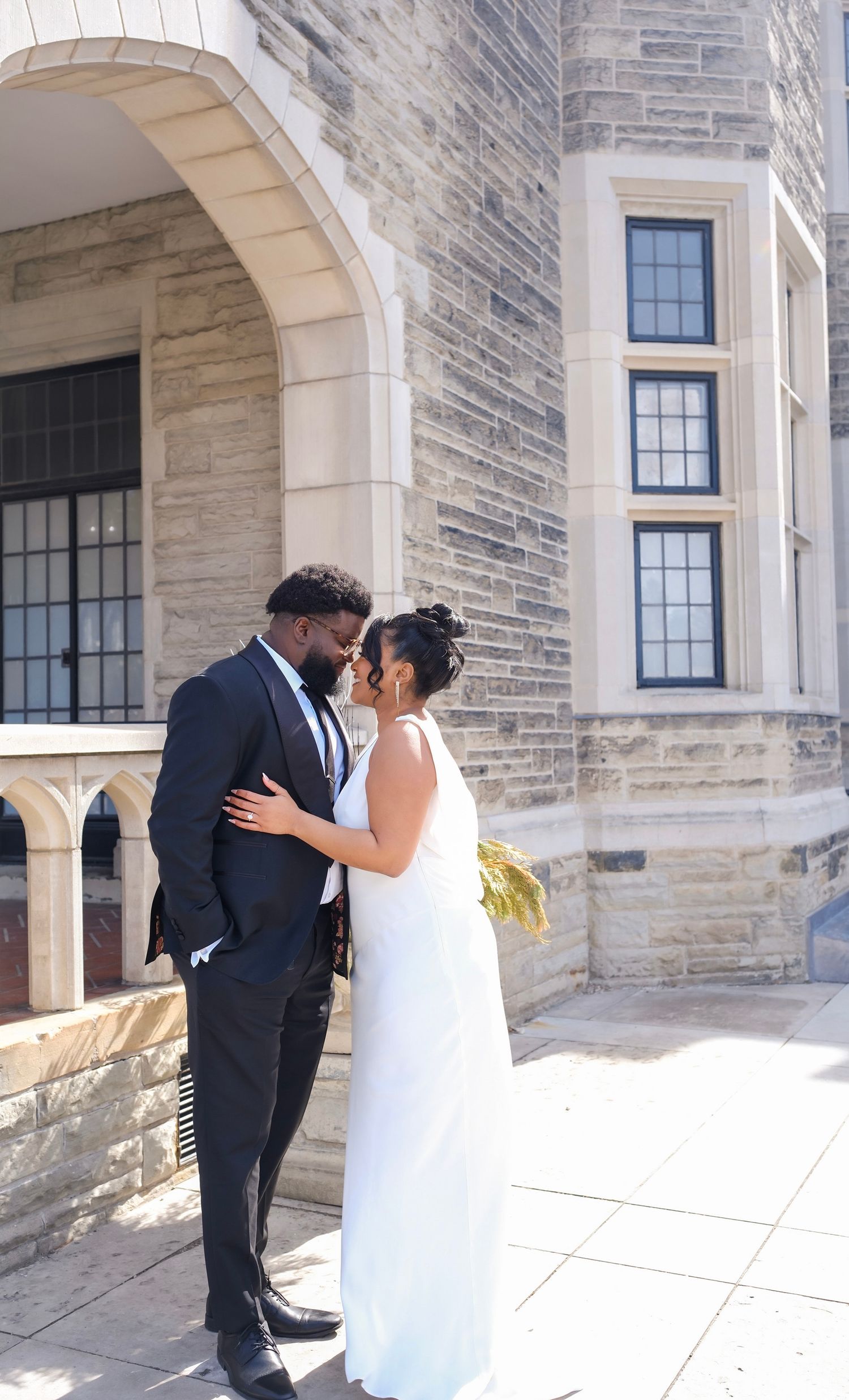 A bride and groom share an intimate moment in front of a stone building with arched windows and architectural details.