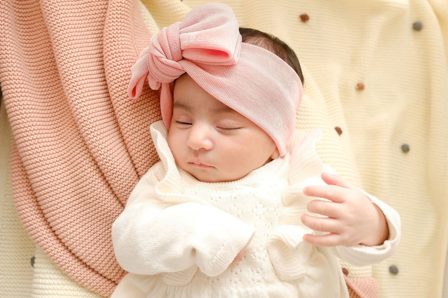Sleeping baby in white outfit wearing pink bow headband resting on pink knit blanket. Toronto, photographer, majestystudios