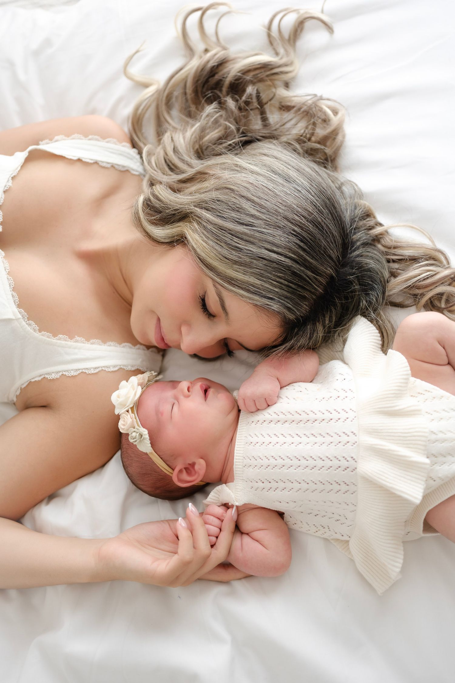 Mother lying on white bed, tenderly nuzzling her newborn baby dressed in white knit outfit with floral headband.