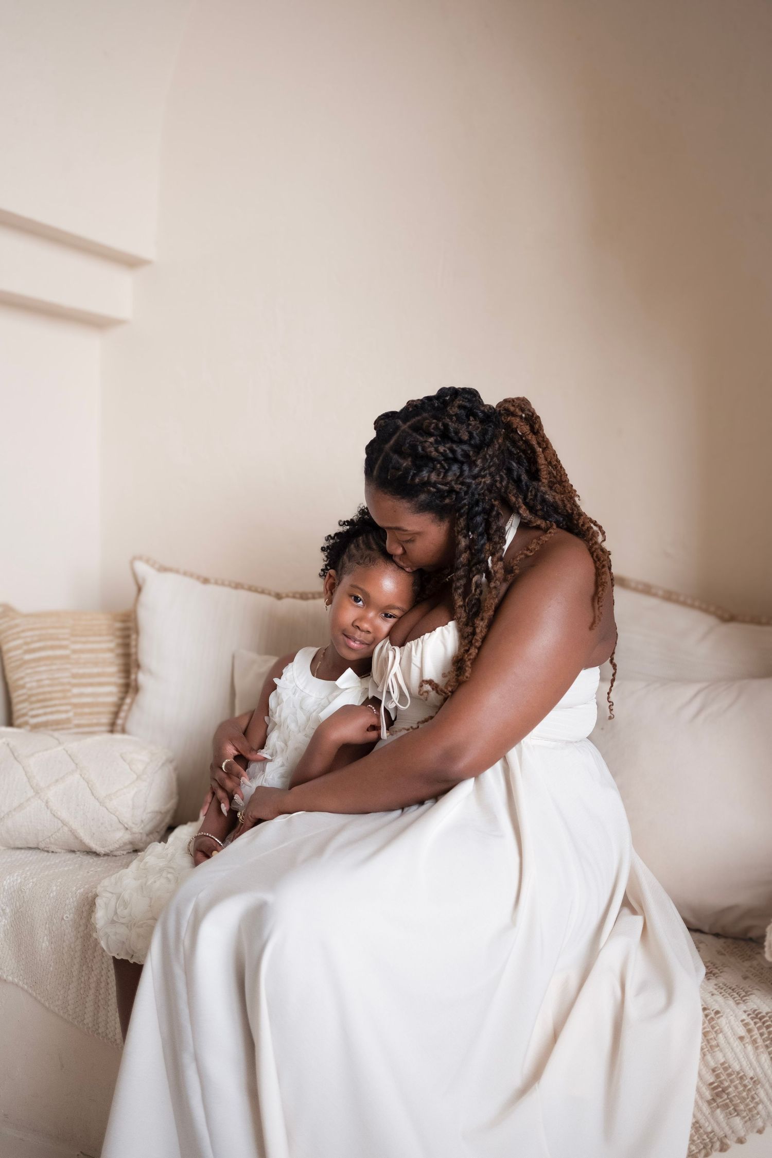 A Black mother in a white dress tenderly embraces her young daughter on a cream sofa.
